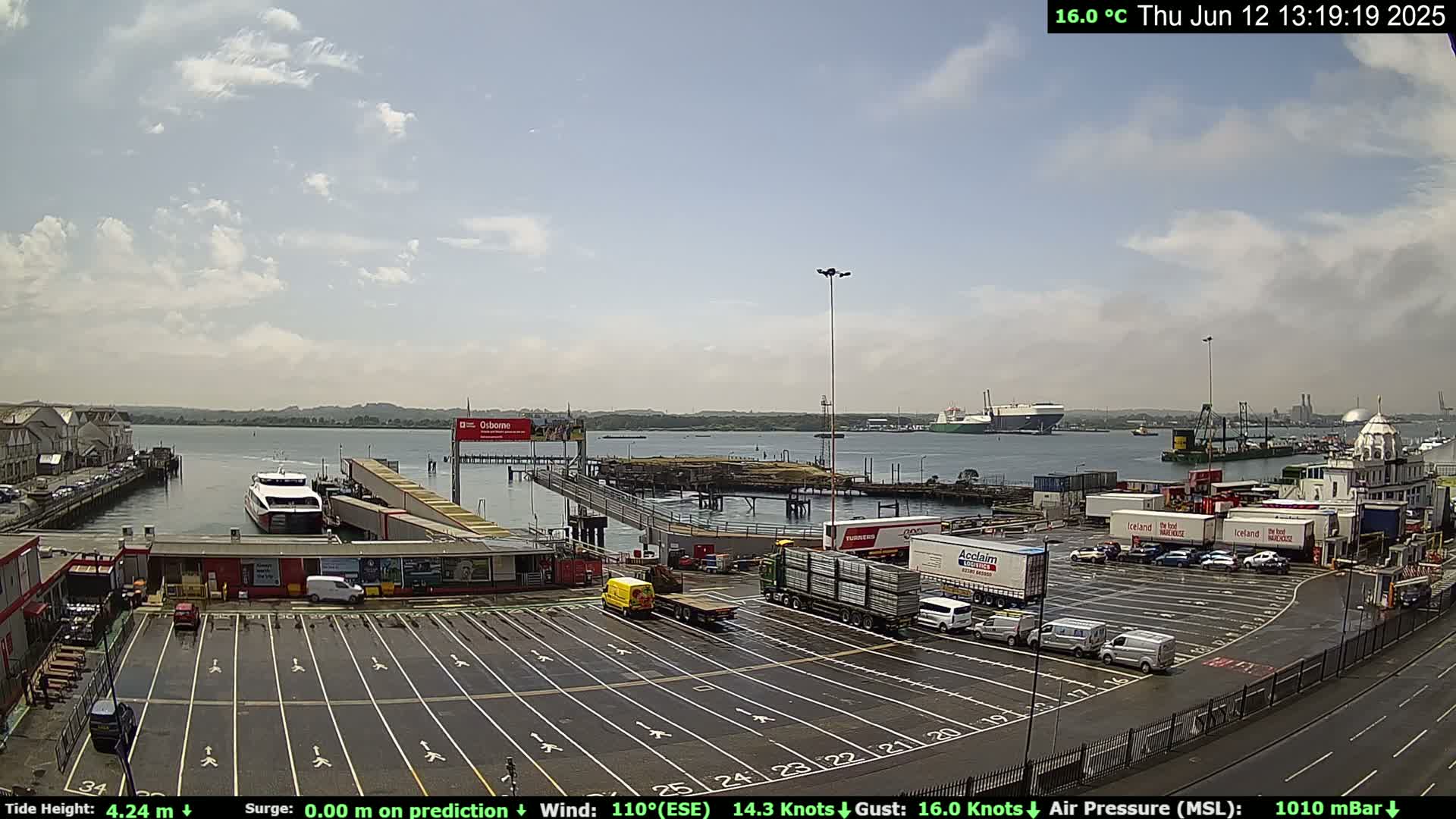 A ferry is docked at a terminal in a harbor on a partly cloudy day, with numerous trucks and cars parked in the surrounding lot.