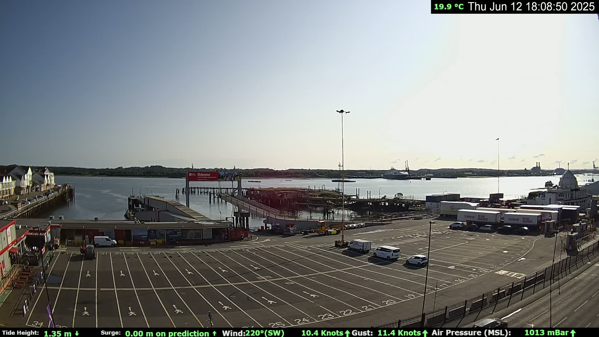 A mostly sunny, outdoor view of a ferry terminal with several vehicles parked in a large lot, and various boats and ships in the water.