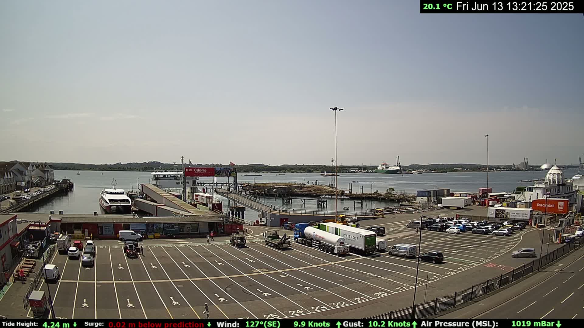 A ferry is docked at a port on a sunny day, with cars parked in a large lot and other vehicles nearby.