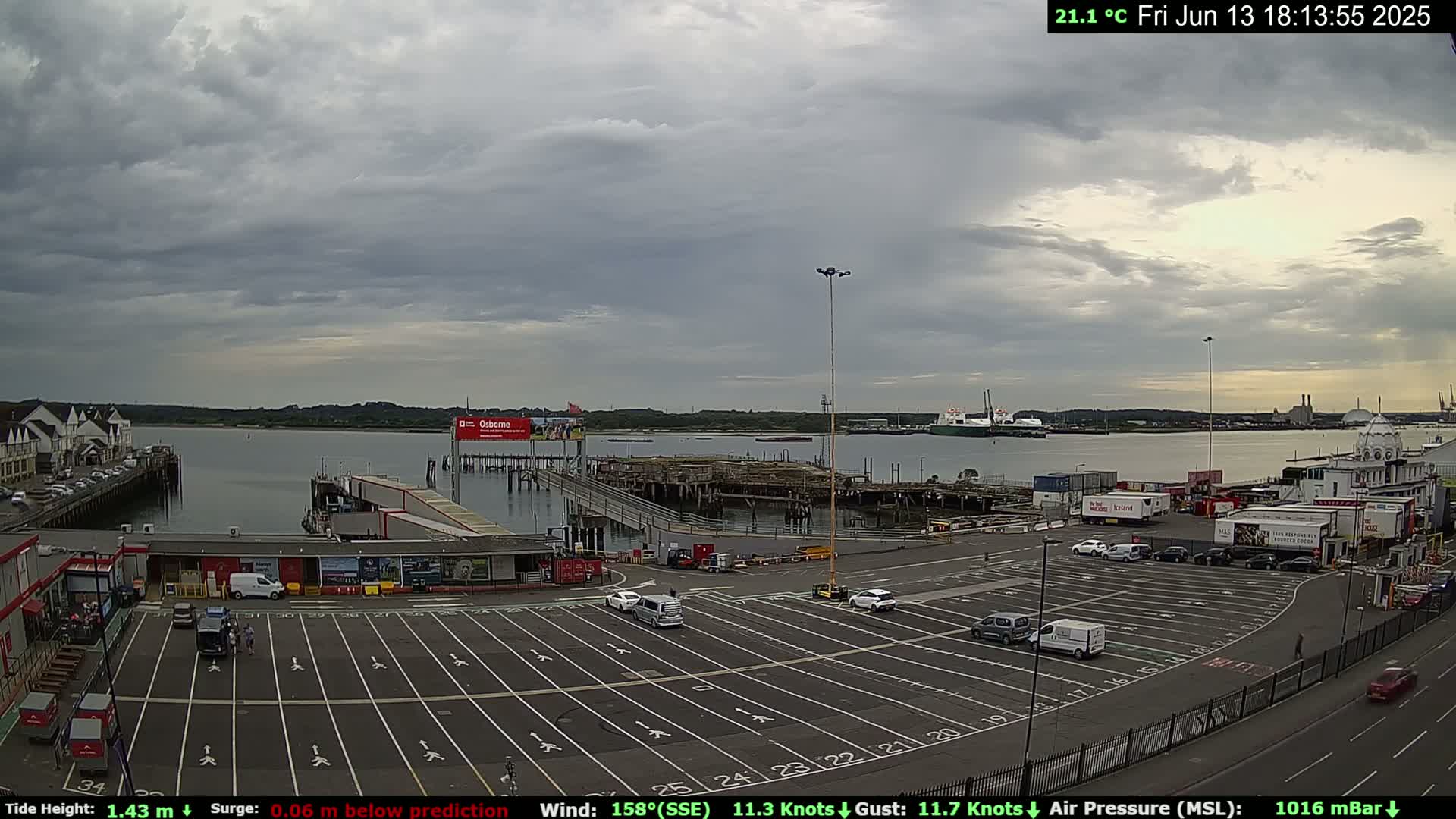 A mostly cloudy day reveals a harbor scene with a large parking lot, several buildings, and docked ships, all under a pale sky.