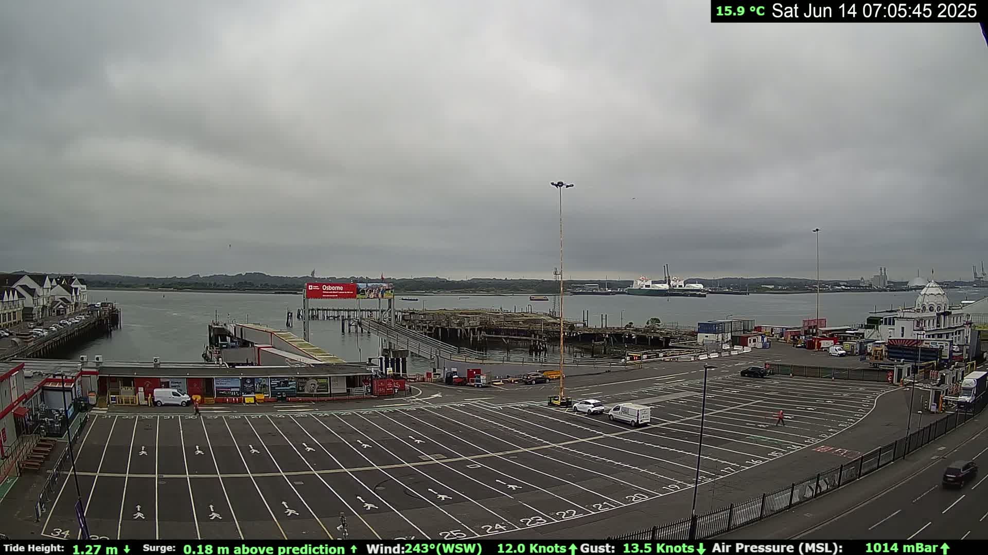 A mostly cloudy day reveals a harbor scene with a large parking lot, several buildings, and docked ships, all under a pale sky.