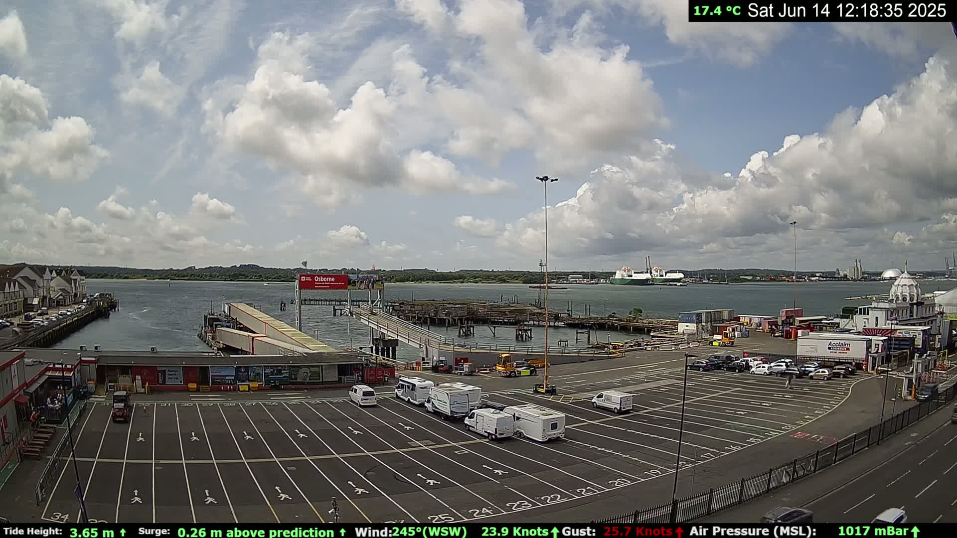 A partly cloudy day shows a ferry terminal with several vehicles parked in the lot, a body of water with ships in the distance, and buildings along the shoreline.