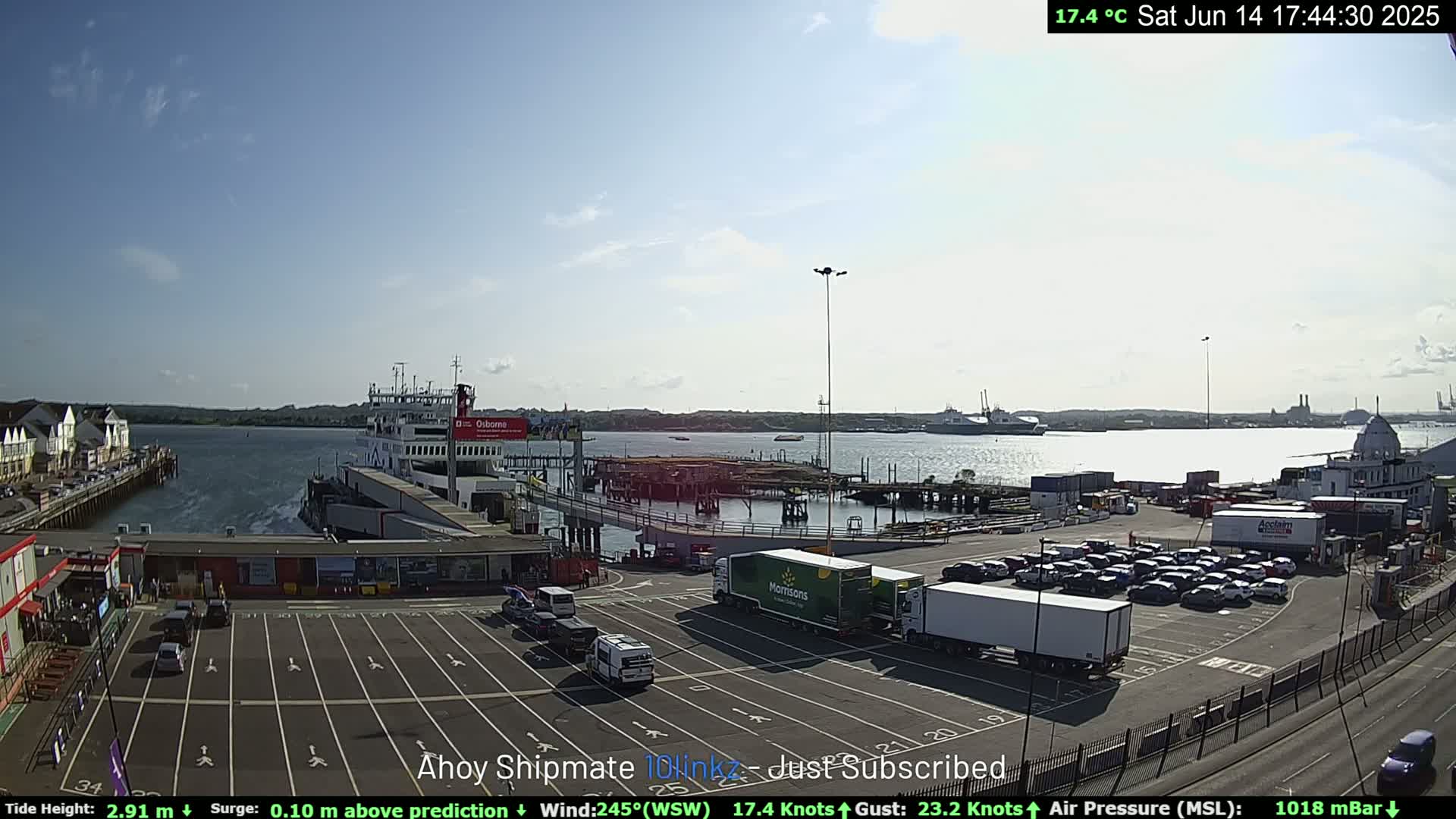 A ferry is docked at a terminal with several cars and trucks in a parking lot, under a mostly sunny sky.