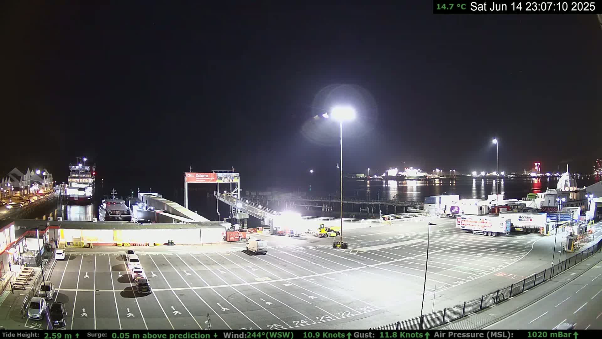 A ferry is docked at a terminal with several cars and trucks in a parking lot, under a mostly sunny sky.