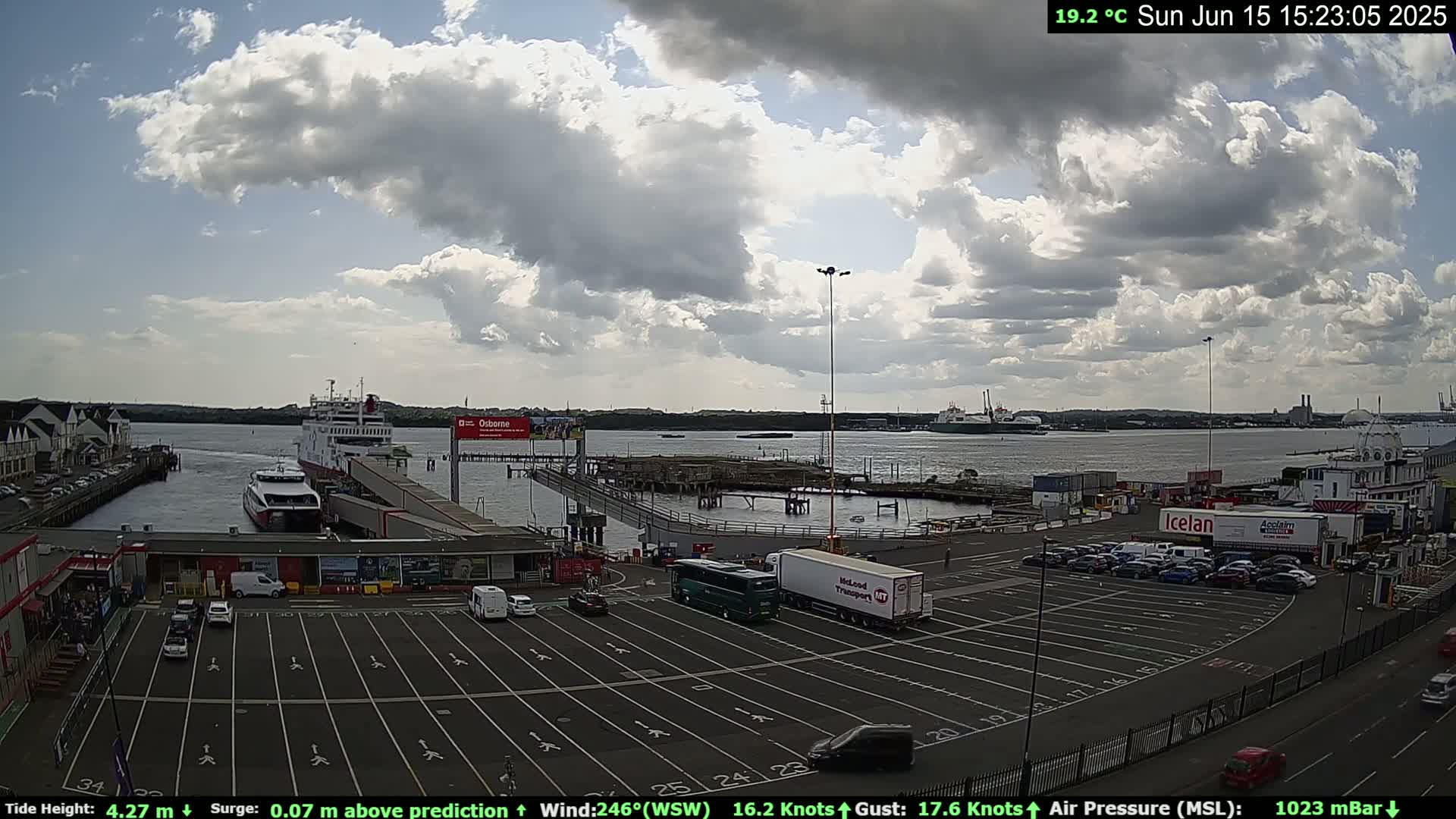 A partly cloudy day at a ferry terminal shows several vehicles parked, with ferries docked and other ships in the distance on calm water.