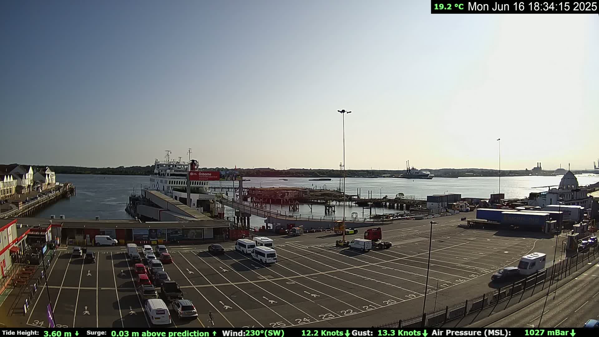 A ferry is docked at a port on a sunny day, with a calm body of water extending to the horizon, and a large parking lot filled with vehicles is visible in the foreground.