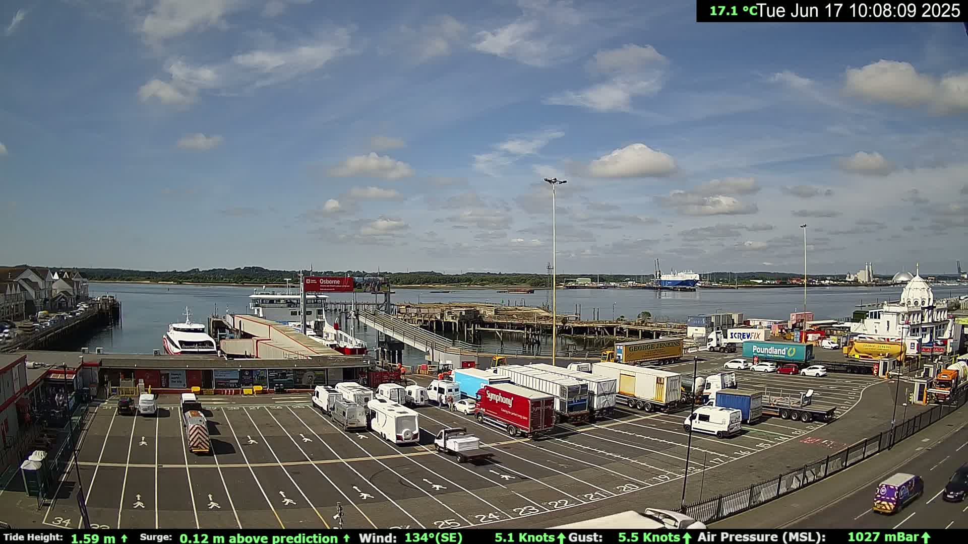 A ferry terminal with numerous vehicles parked in a lot, a ferry docked at the pier, and calm waters under a partly cloudy sky.