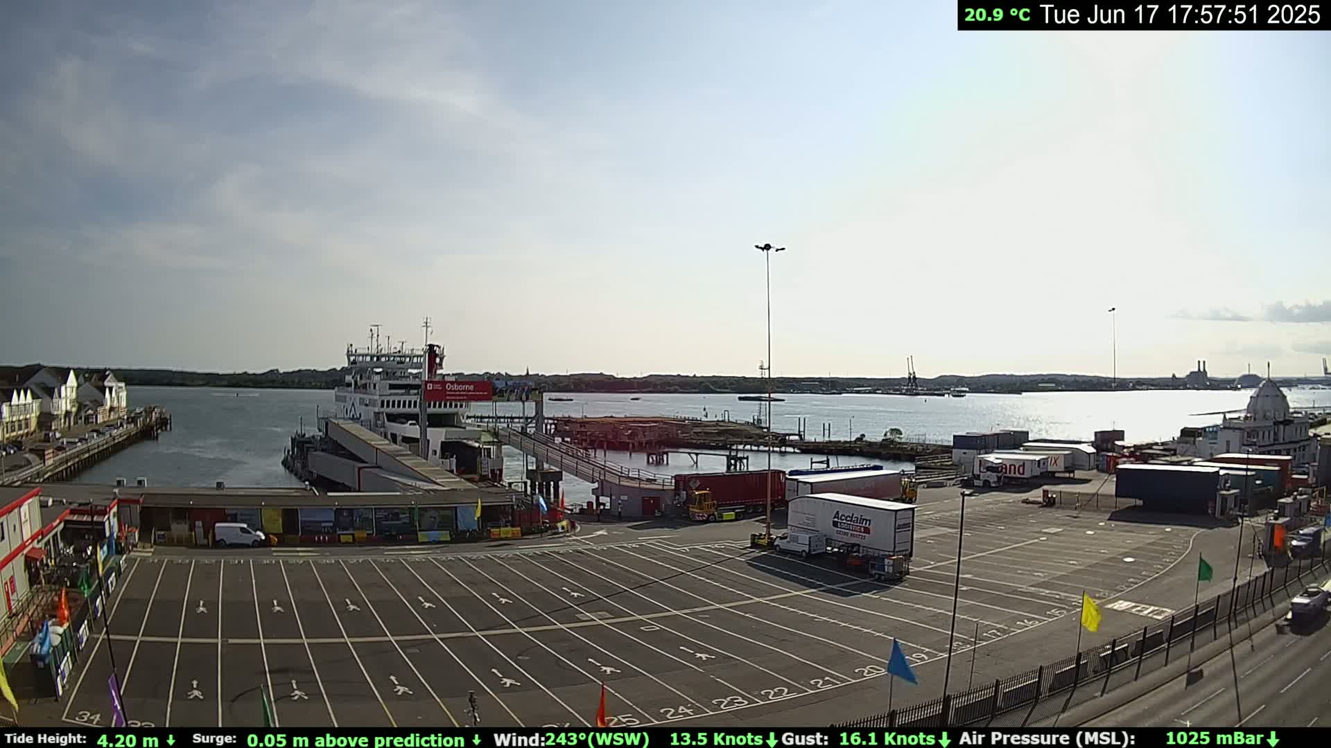 A ferry is docked at a terminal, with numerous trucks and cars in a large parking lot, under a mostly sunny sky.