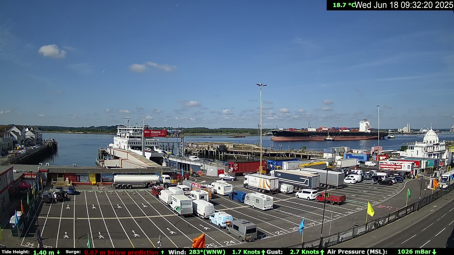 A ferry is docked at a terminal, with a large cargo ship in the background, on a sunny day with a few clouds in the sky.