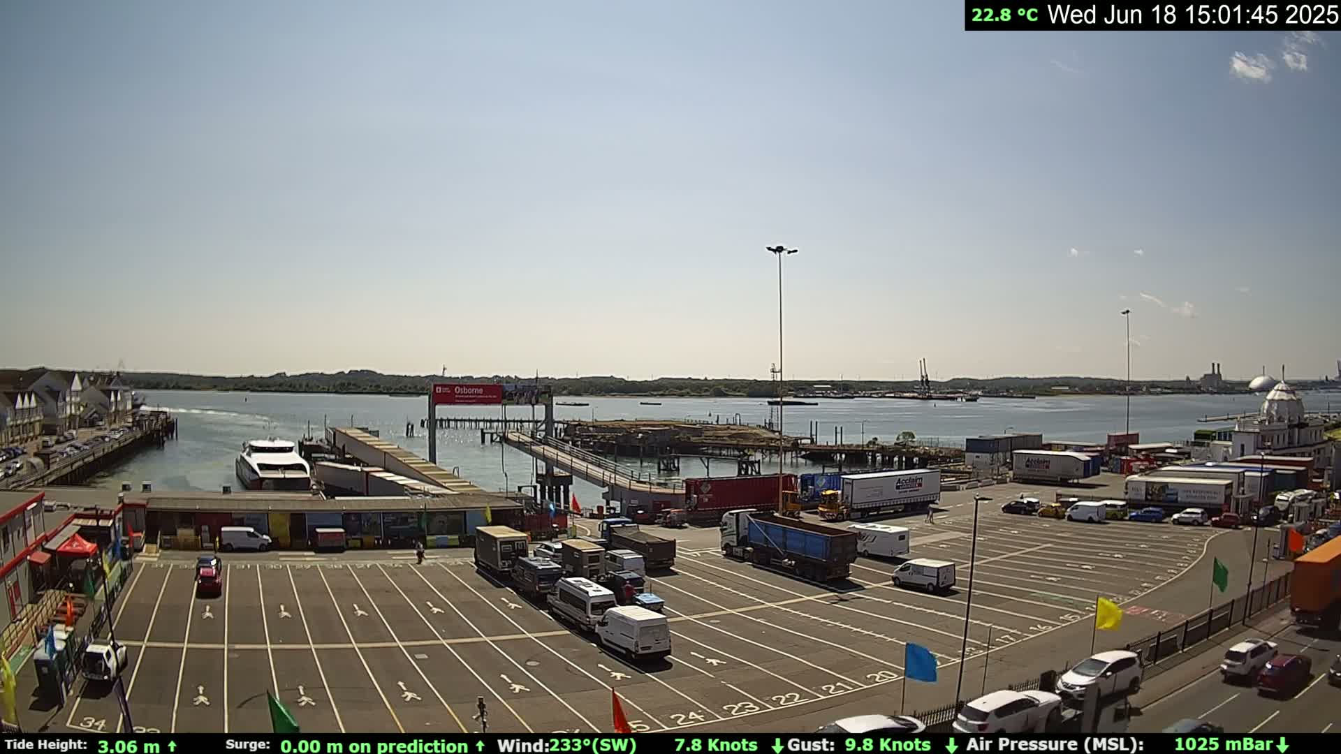 A ferry is docked at a terminal in a sunny harbor with numerous vehicles parked in a lot nearby.