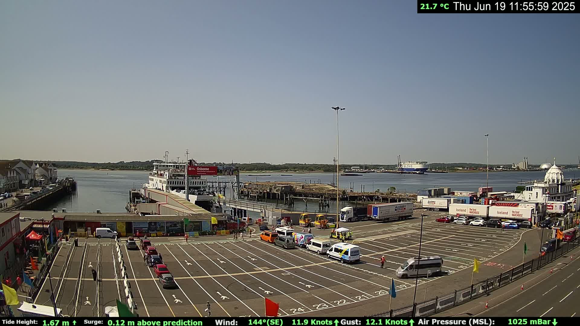 A ferry is docked at a terminal on a sunny day, with numerous cars and trucks parked in the adjacent lot.