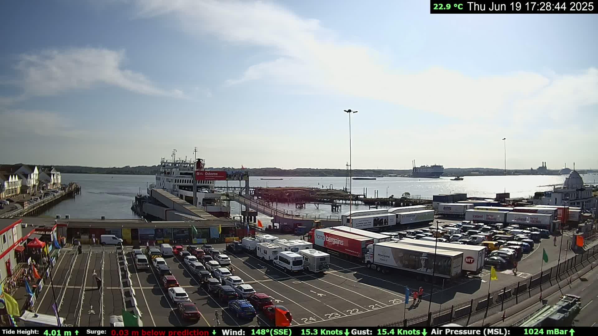 A ferry is docked at a terminal, with numerous cars and trucks parked nearby under a partly cloudy sky.