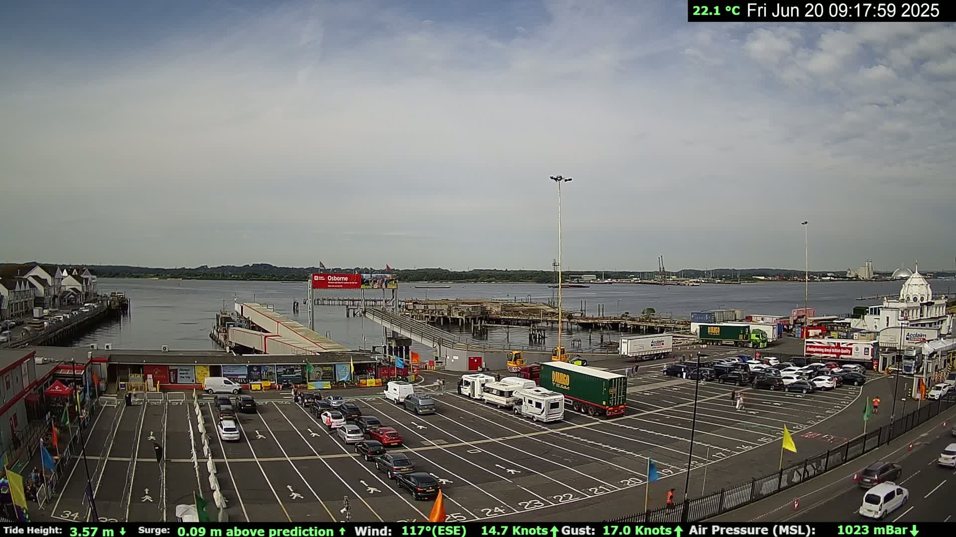 A mostly sunny day at a ferry terminal shows a large parking lot filled with cars and RVs, with a body of water and buildings in the background.
