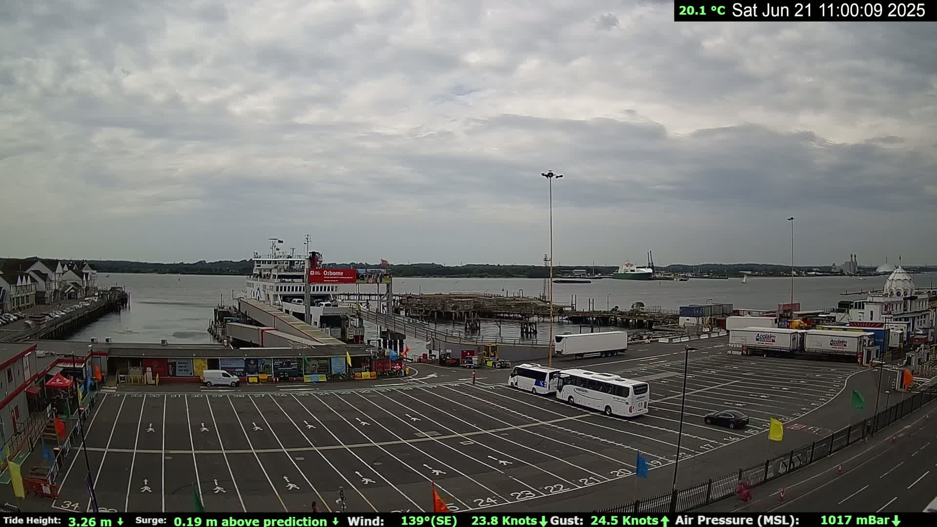A ferry is docked at a port under a cloudy sky, with several buses and cars parked in the adjacent lot.
