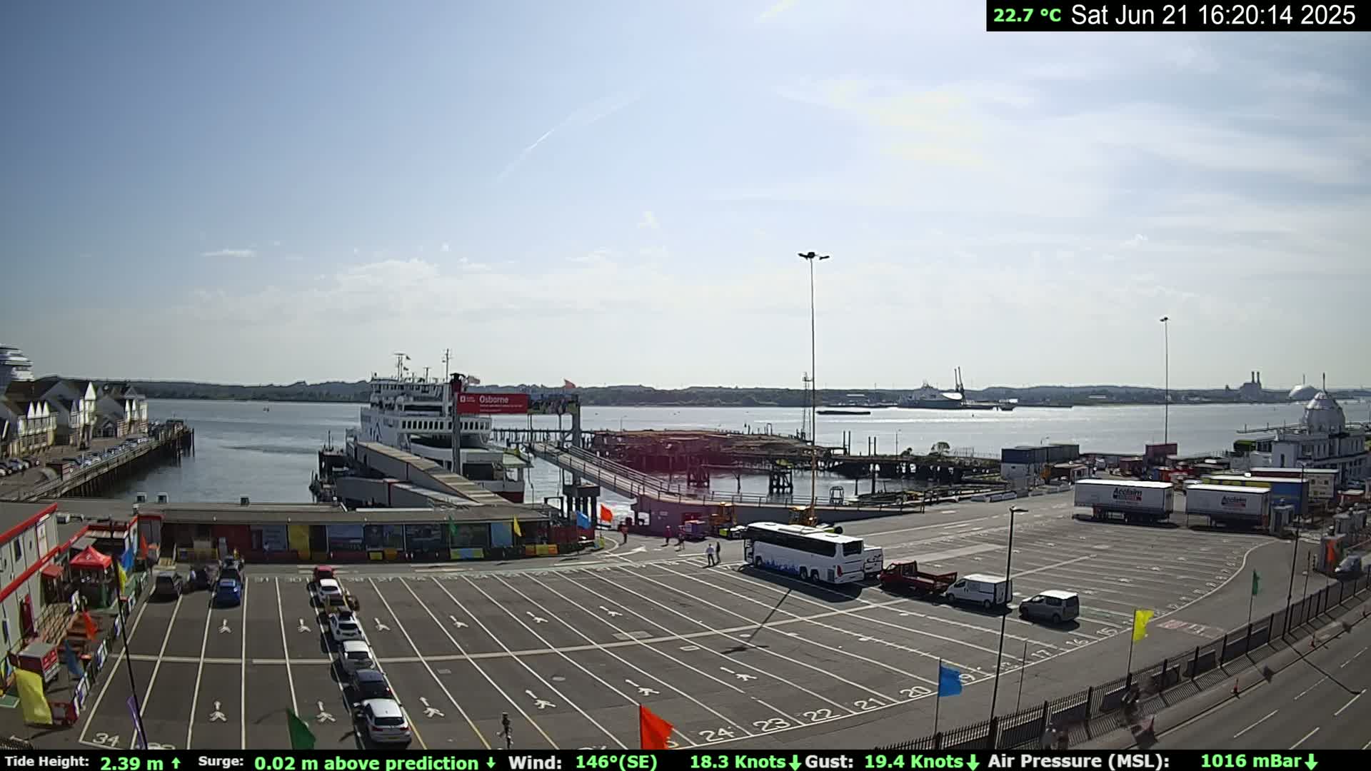 A ferry is docked at a port on a sunny day with a partly cloudy sky, and several vehicles are parked in the surrounding lot.