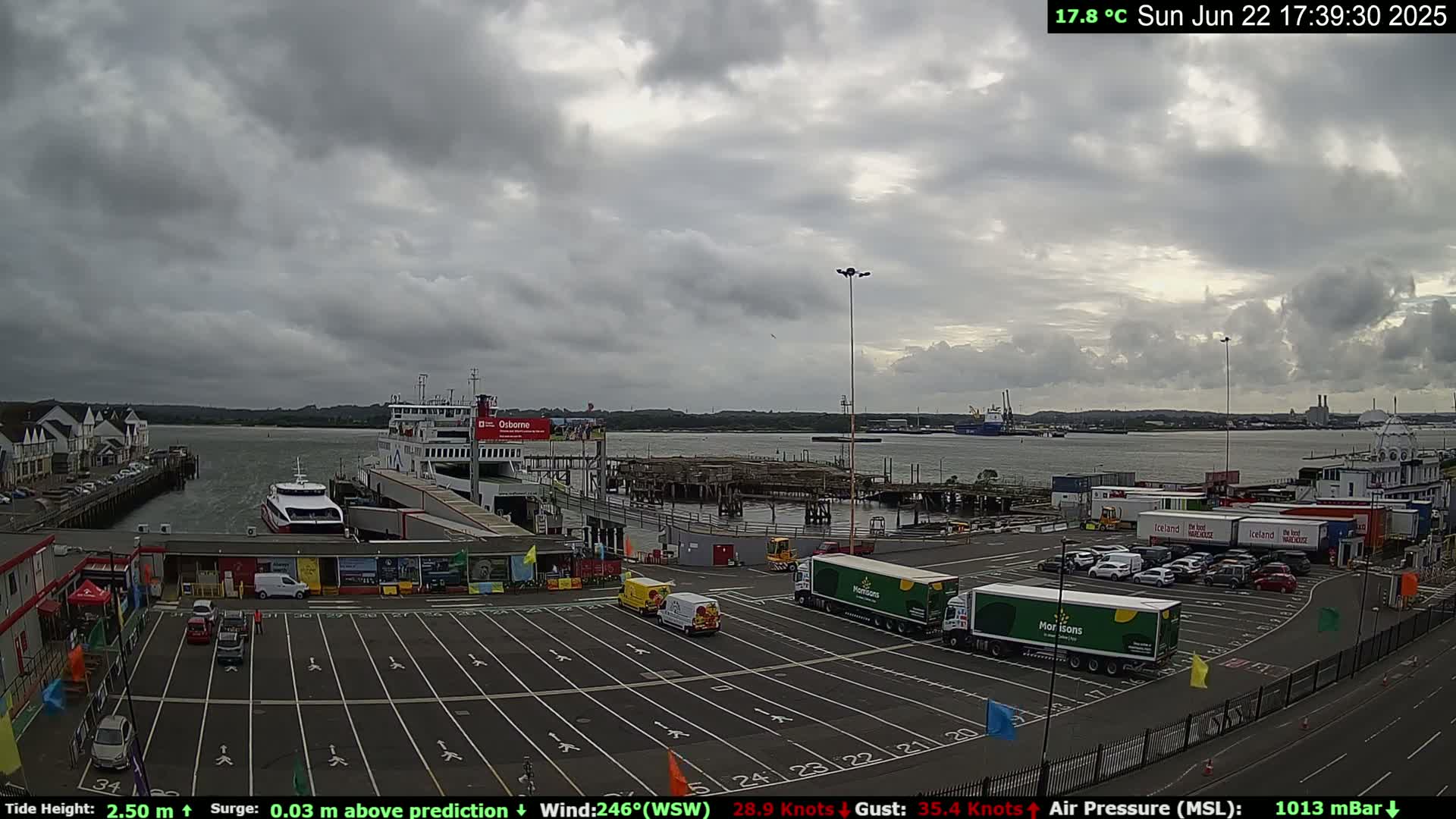 A ferry terminal with several ferries and numerous delivery trucks parked in a large lot under an overcast sky.