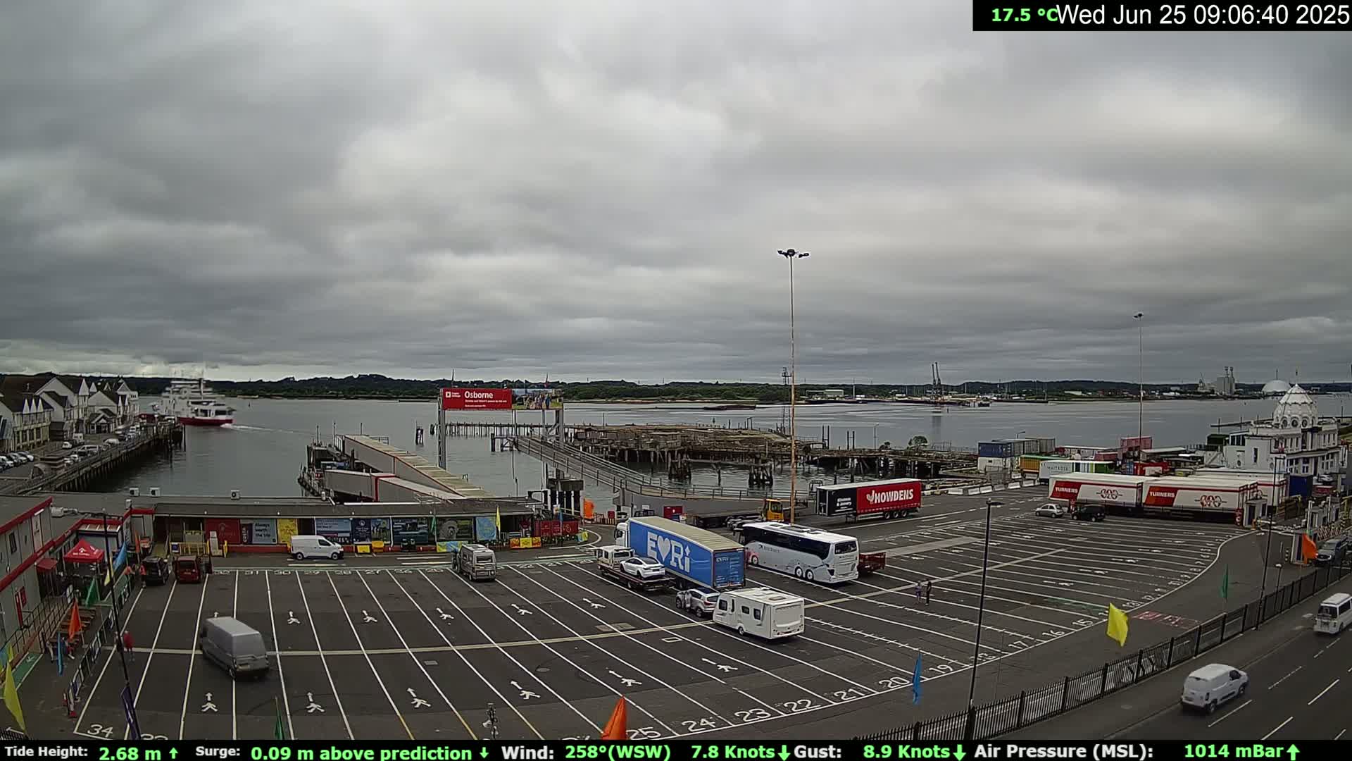 A ferry departs a harbor under an overcast sky, with numerous vehicles parked in a lot near the terminal.