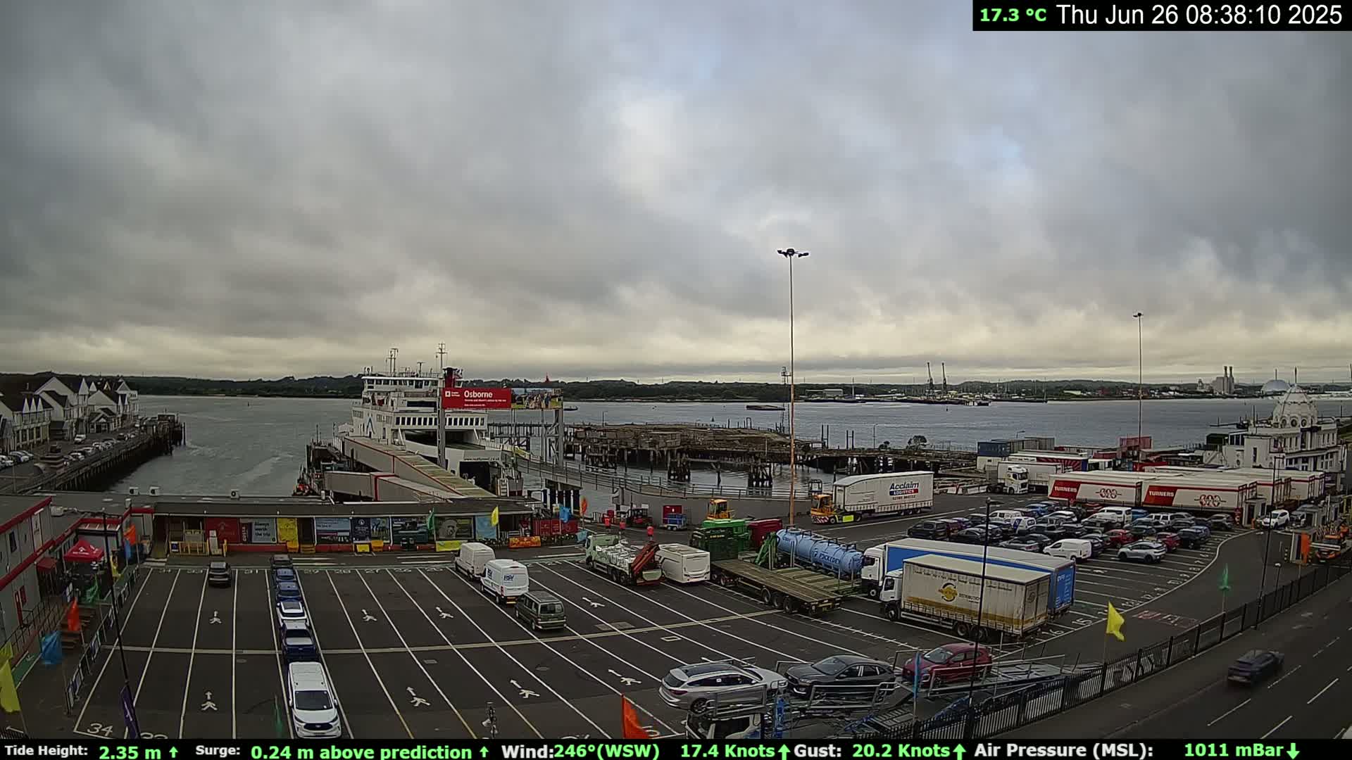 A ferry is docked at a terminal with overcast skies, and numerous vehicles are parked in the lot.