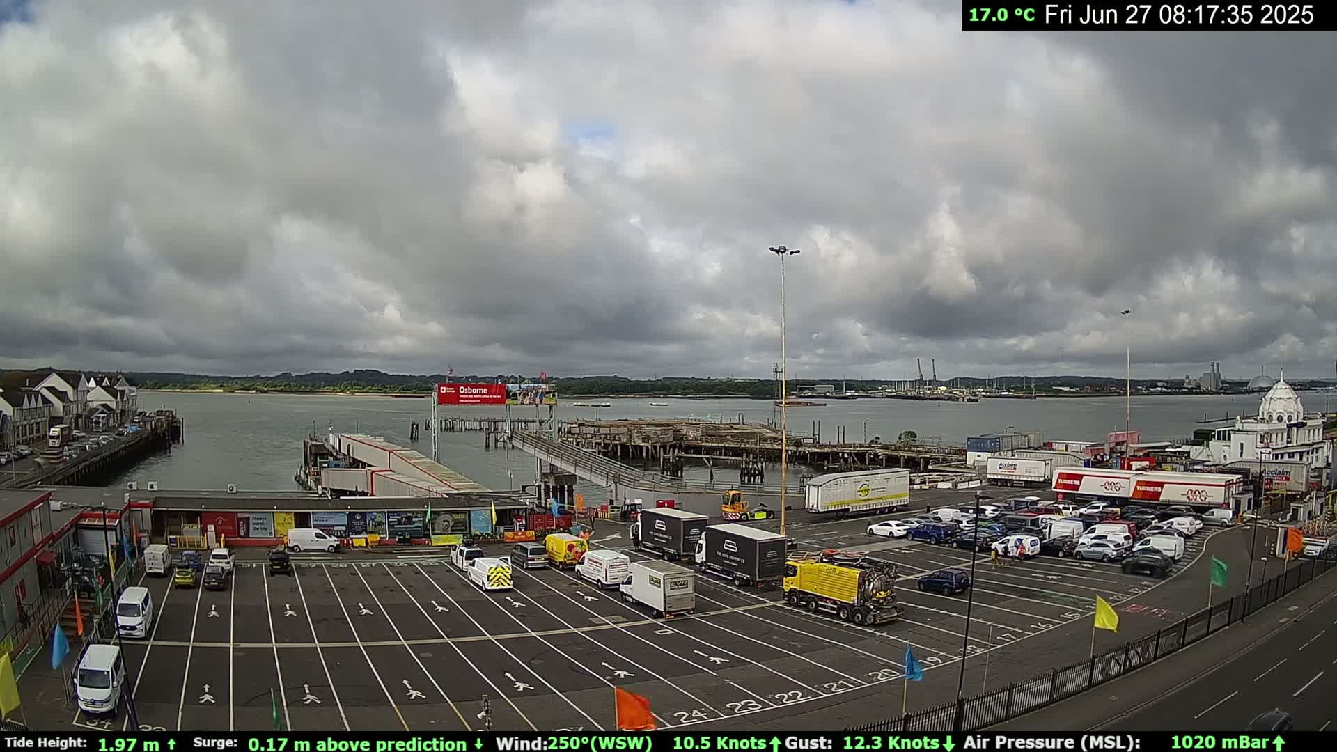 A ferry terminal parking lot with numerous vehicles, under a mostly cloudy sky, overlooks a calm body of water.