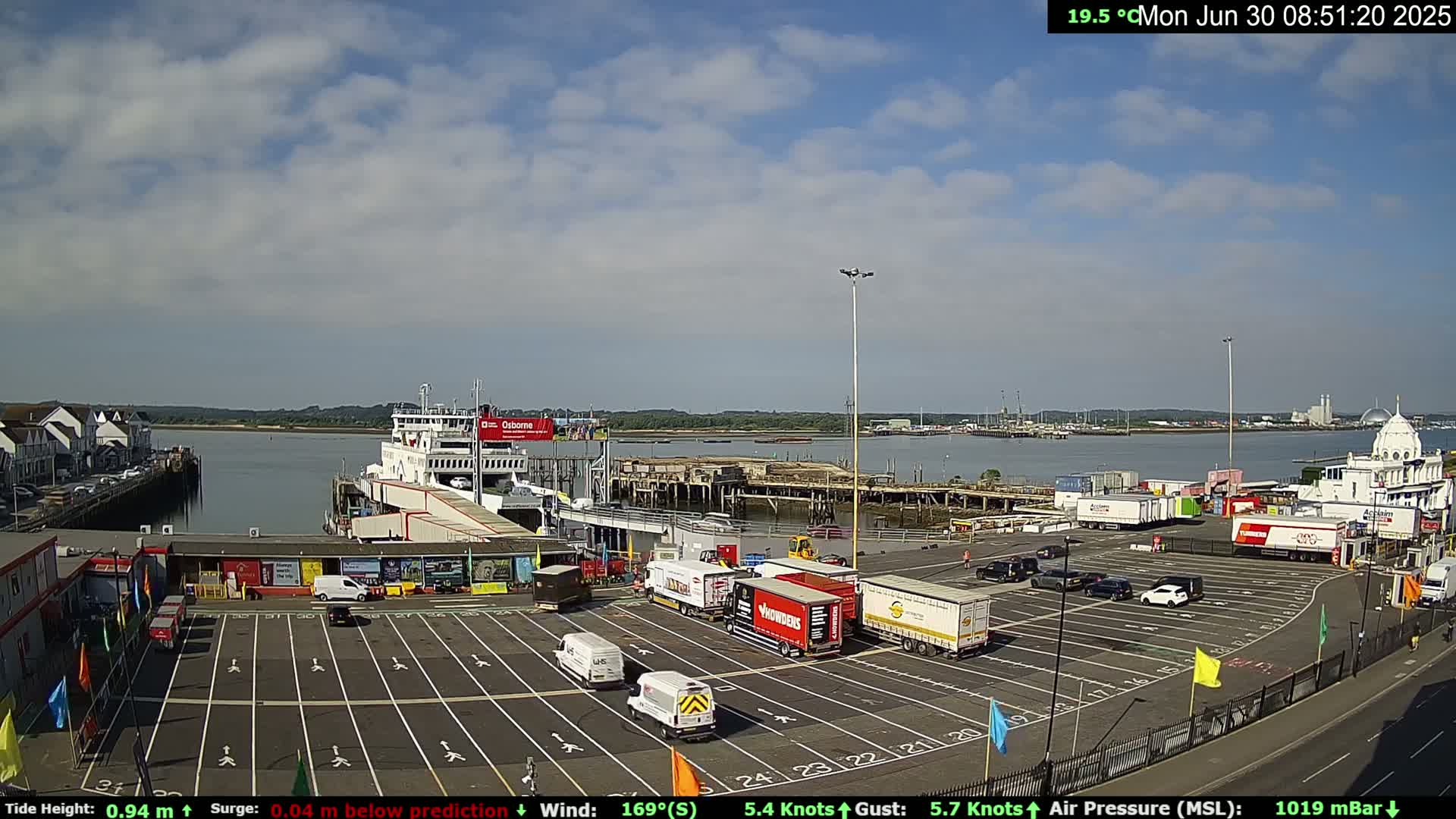 A ferry is docked at a port on a partly cloudy day, with numerous trucks and cars parked in the surrounding area.