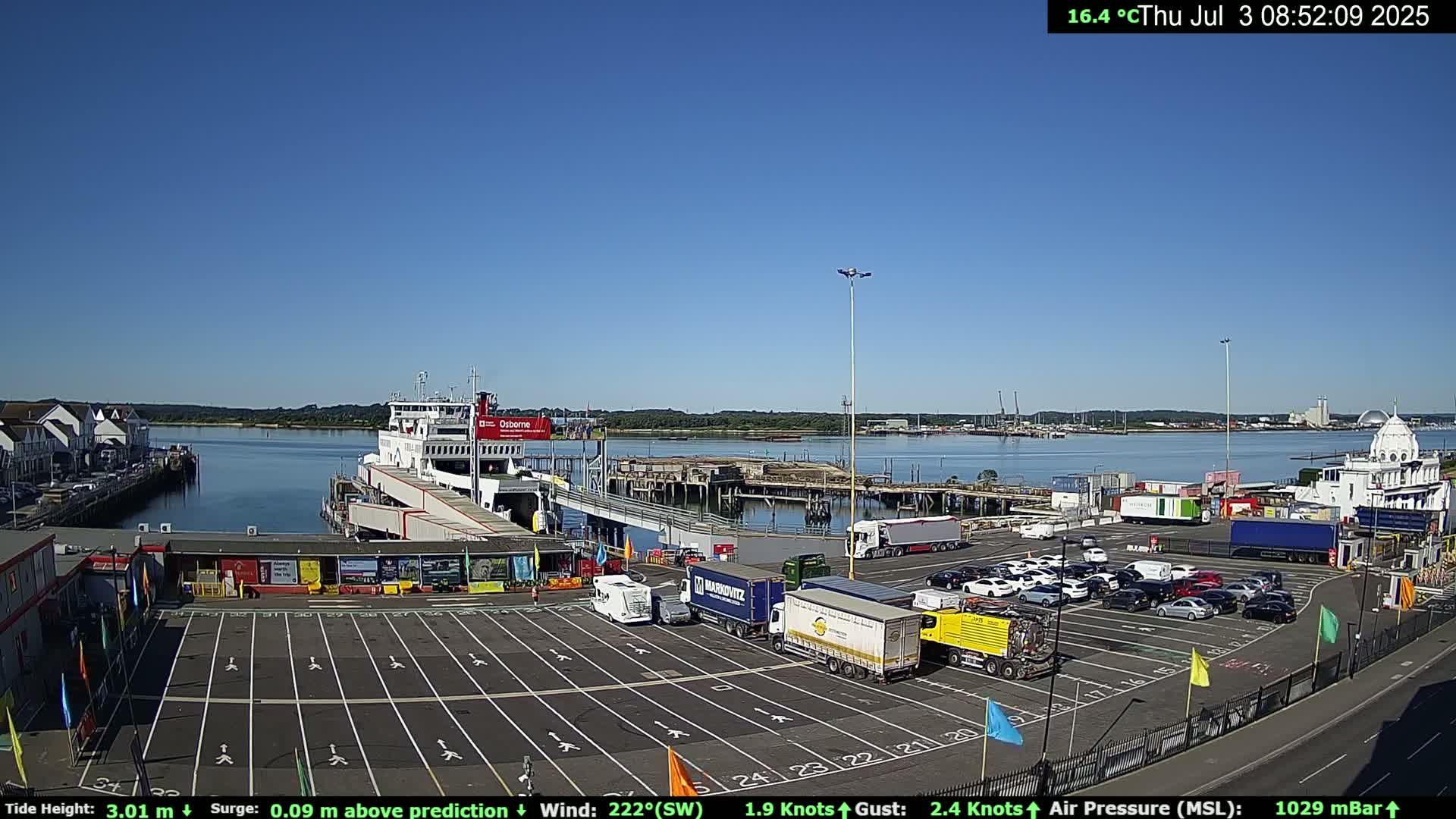 A ferry is docked at a terminal on a sunny day, with numerous cars and trucks parked in the surrounding lot and calm water visible in the background.