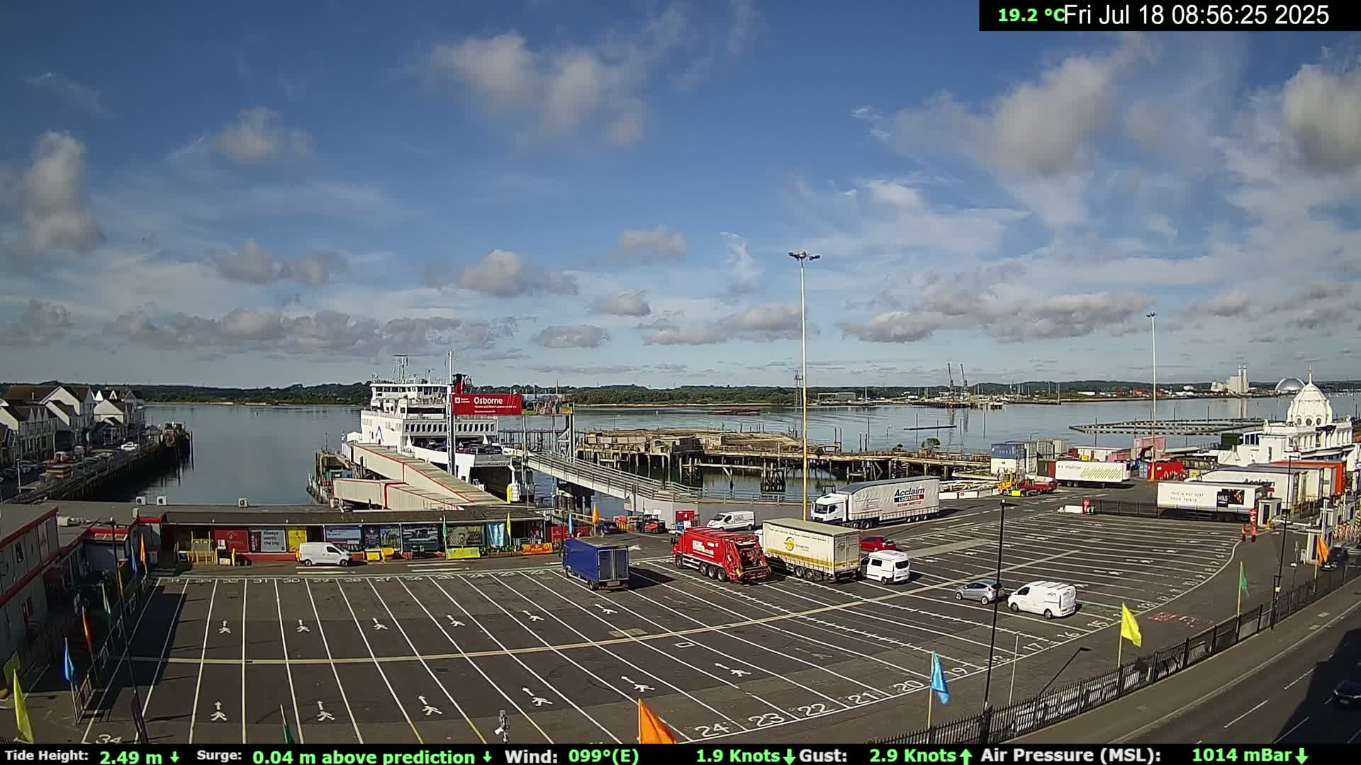 A ferry is docked at a terminal with a mostly sunny sky and a few clouds, and numerous trucks and cars are parked in the adjacent lot.