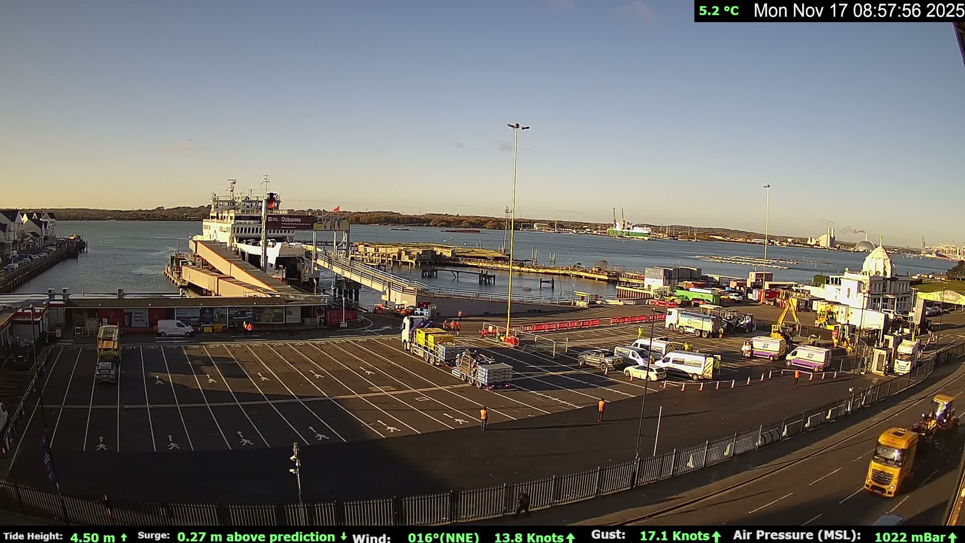 A sunny morning reveals a busy ferry terminal with a large white ferry docked at the pier, numerous trucks and vans on the expansive tarmac, and calm harbor waters stretching towards distant industrial structures under a clear blue sky.