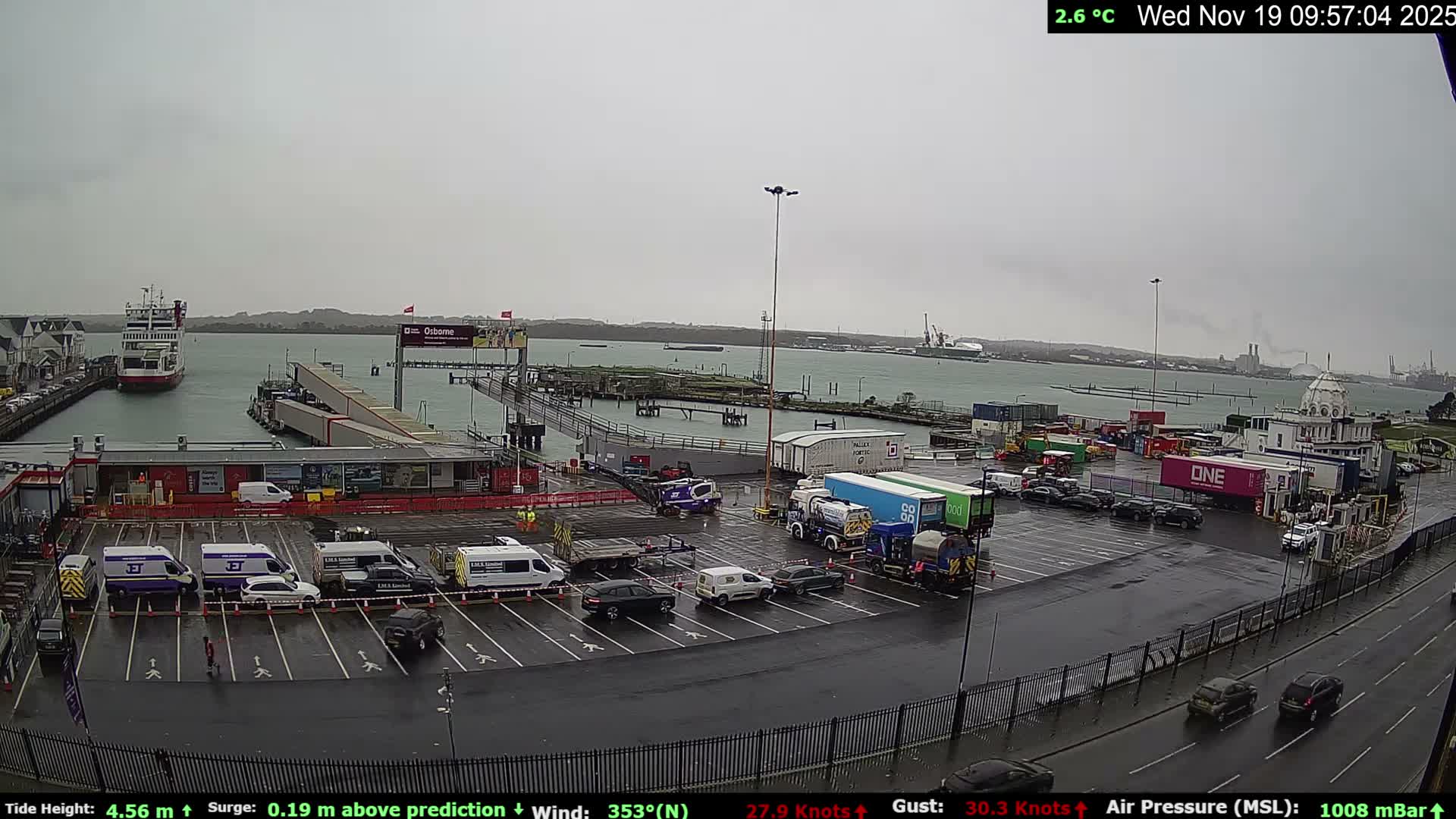 A busy ferry terminal and port with a ferry docked, numerous vehicles in a wet parking area, and distant industrial structures along a bay is captured under a gloomy, overcast sky.