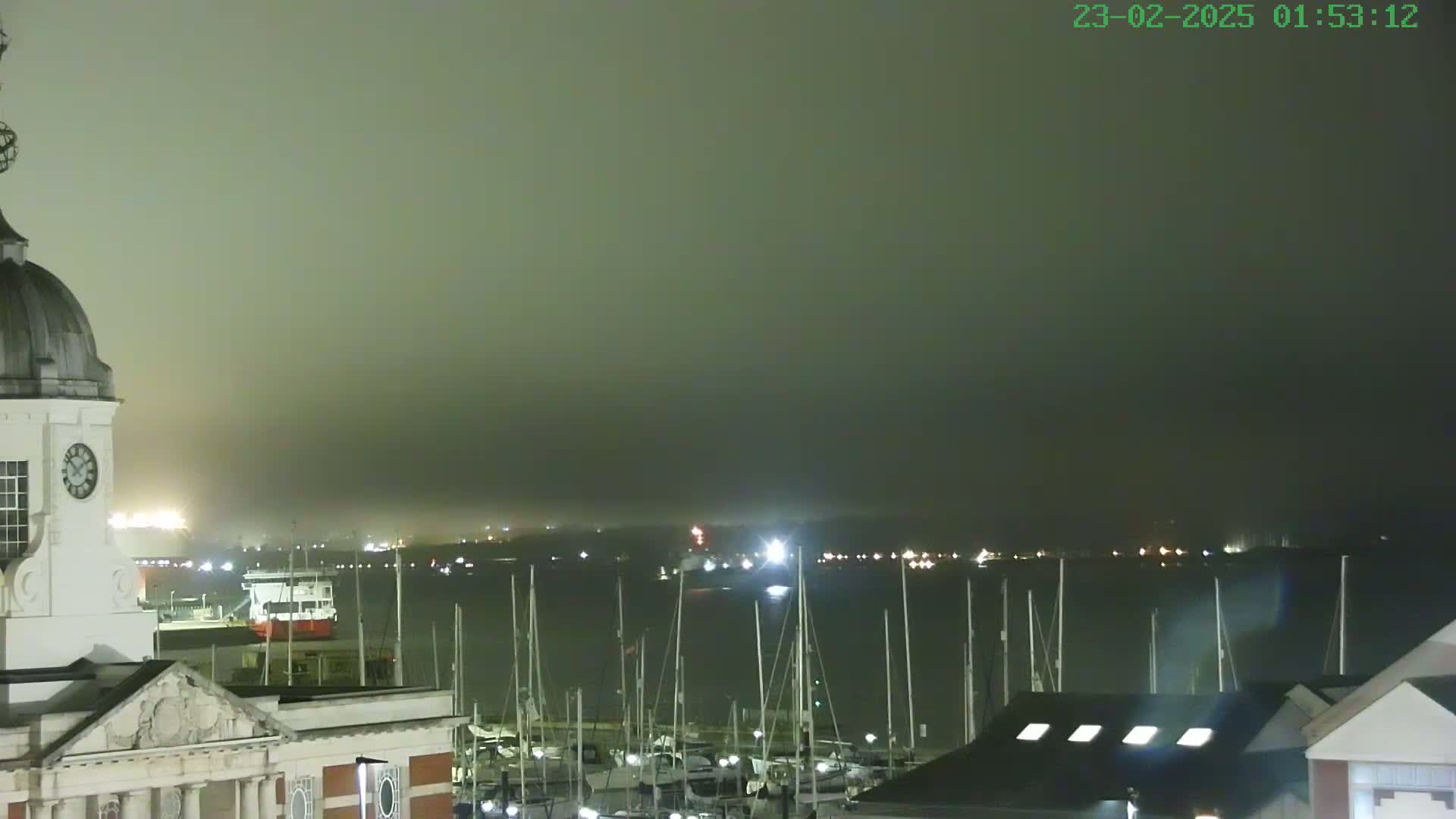 A foggy night scene shows a harbor filled with docked sailboats and a large building with a clock tower in the foreground.