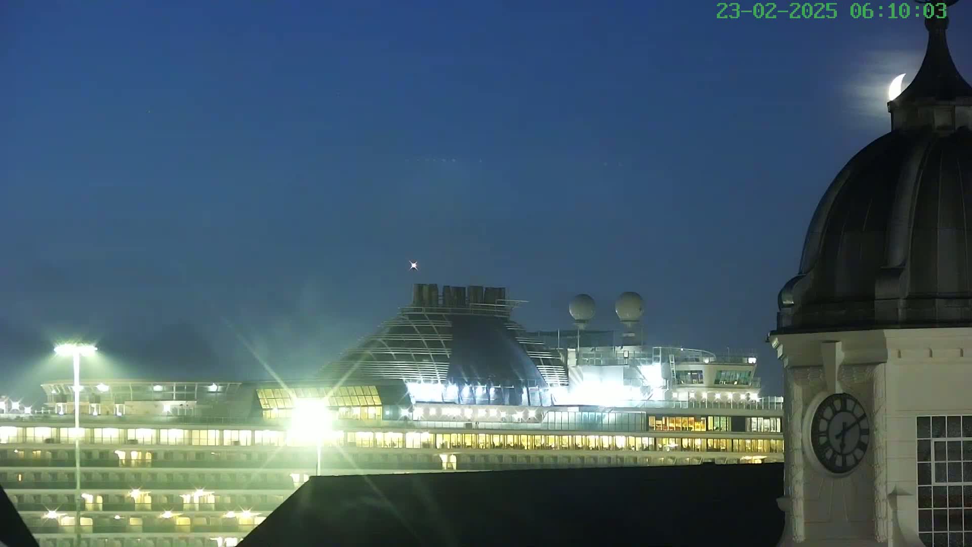 A brightly lit cruise ship is docked at night under a twilight sky with a crescent moon visible, partially obscured by a building with a clock tower.