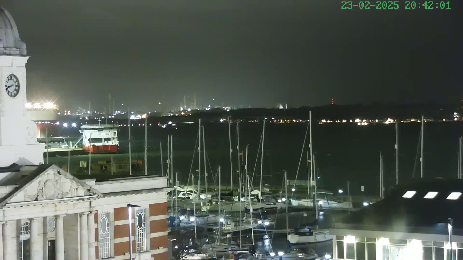 A nighttime view of a harbor with numerous sailboats docked, a large ship in the background, and a clock tower on a brick building in the foreground.