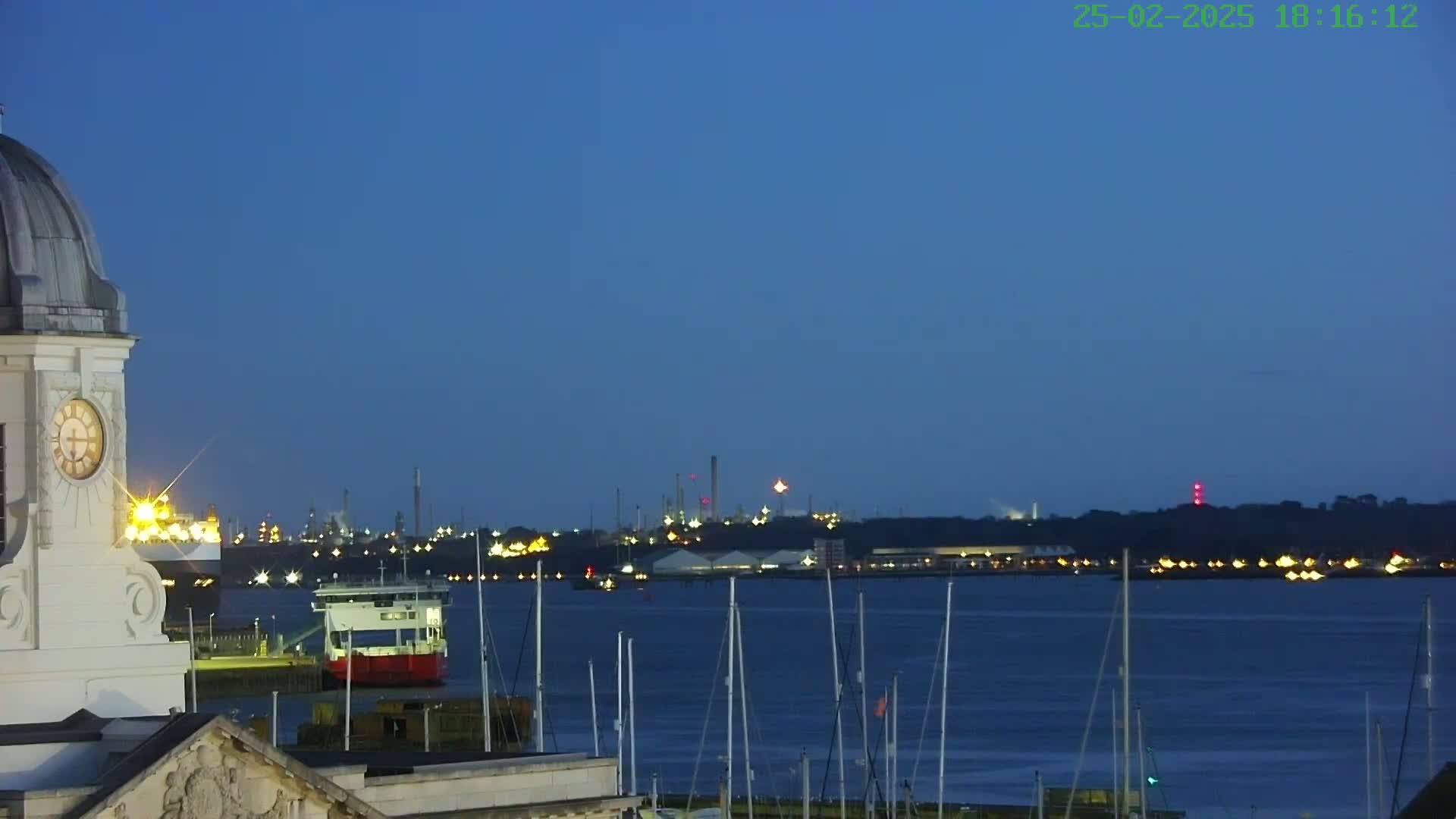 A clock tower overlooks a harbor at twilight, with numerous sailboats moored and an industrial area faintly lit in the distance.