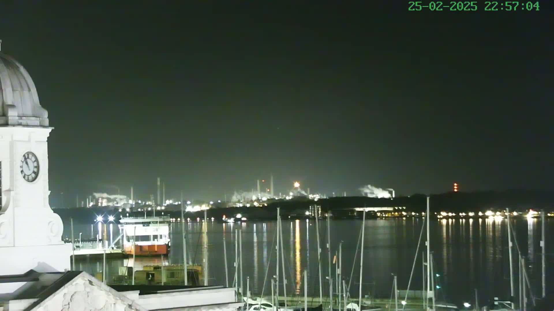 A nighttime view from a clock tower shows a calm body of water with numerous masts of sailboats and a distant industrial complex illuminated under a dark sky.