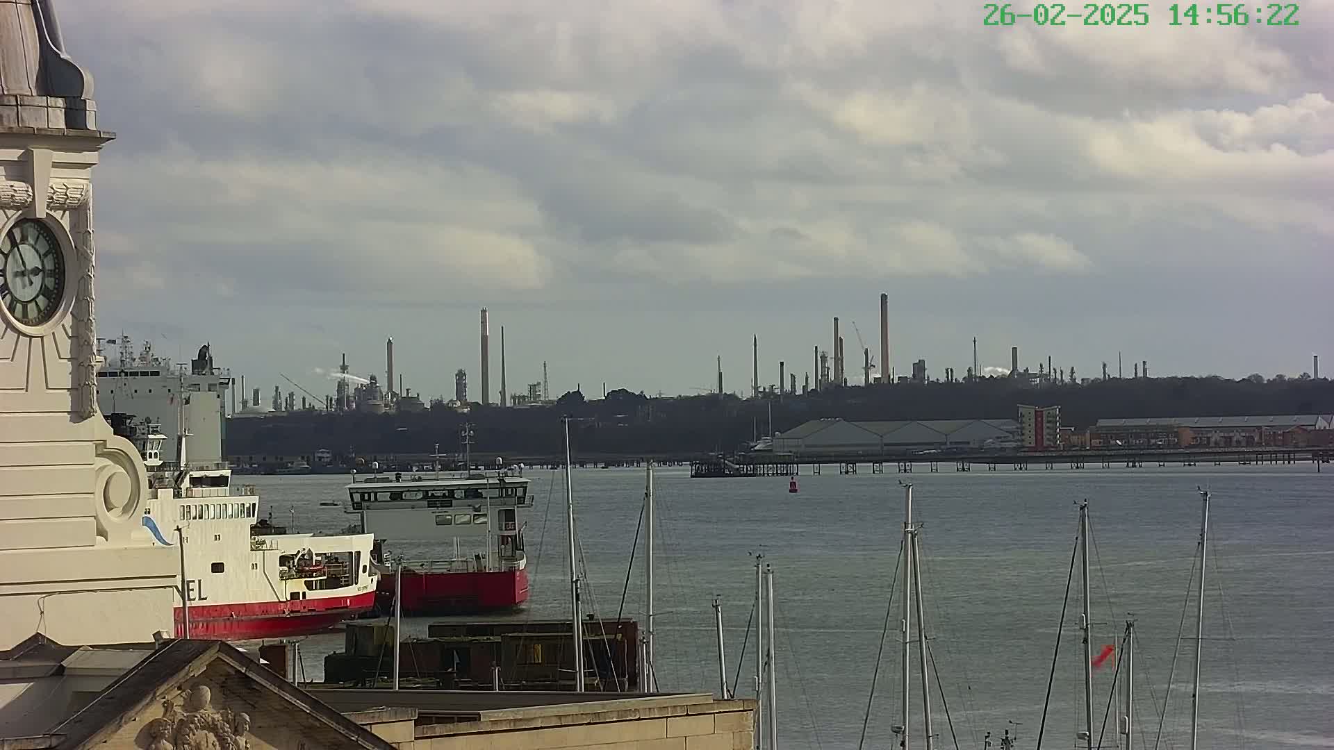 A clock tower overlooks a harbor with several ships, and in the distance, an industrial refinery is visible under a cloudy sky.