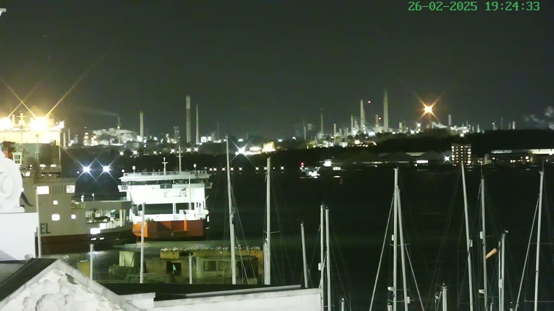At night, a harbor with docked ships and boats is seen in the foreground, with an illuminated industrial plant in the background under a clear, dark sky.