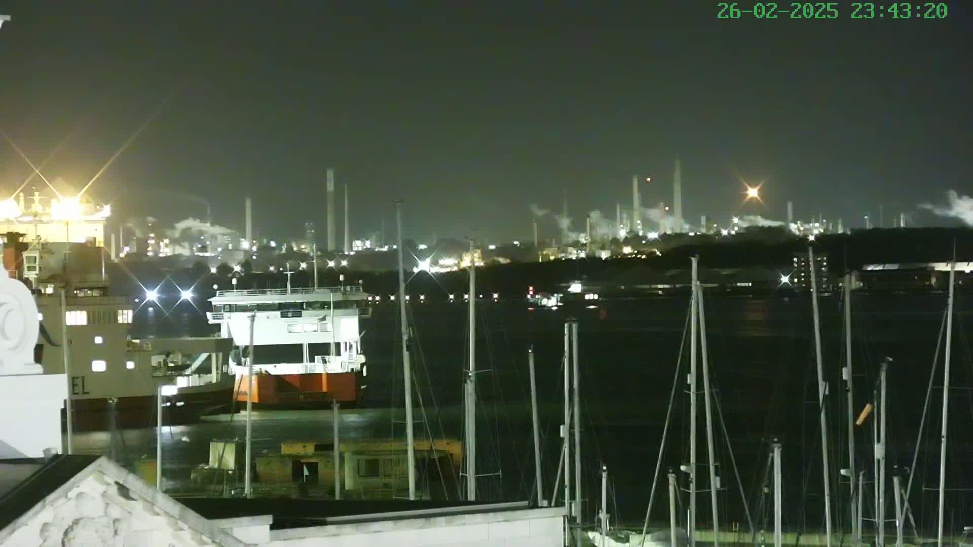 At night, a harbor with docked ships and boats is seen in front of an illuminated industrial complex emitting smoke under a dark sky.