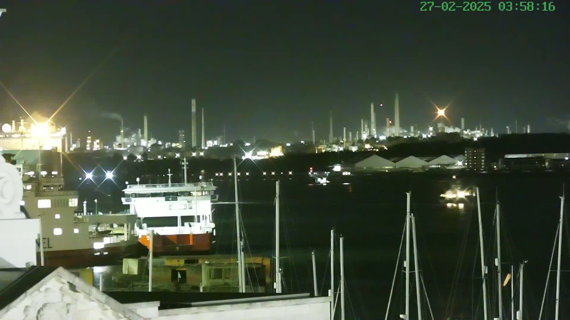 A nighttime view of a harbor with docked ships and boats in front of an illuminated industrial plant under a clear sky.