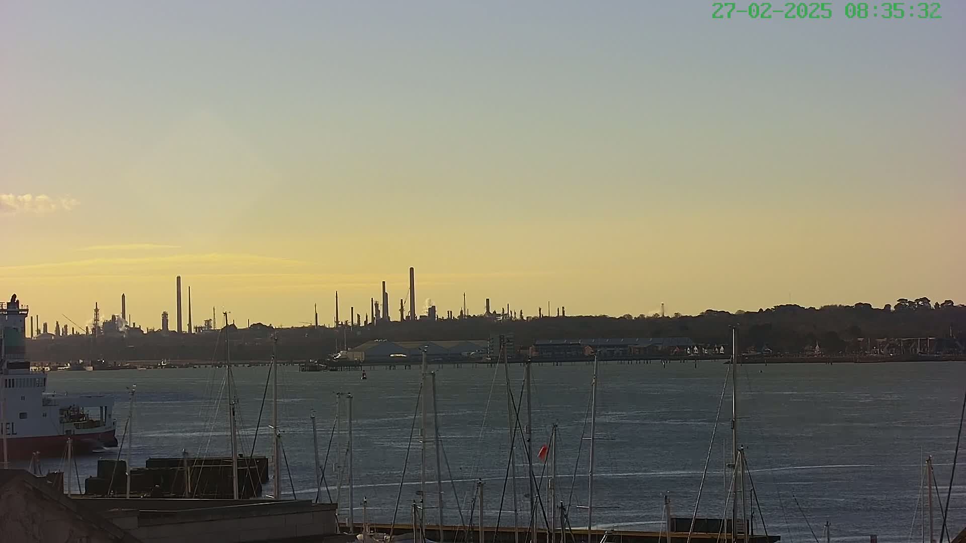 A calm body of water with numerous sailboats in the foreground, a large industrial plant silhouetted against a pale yellow sunset in the background.