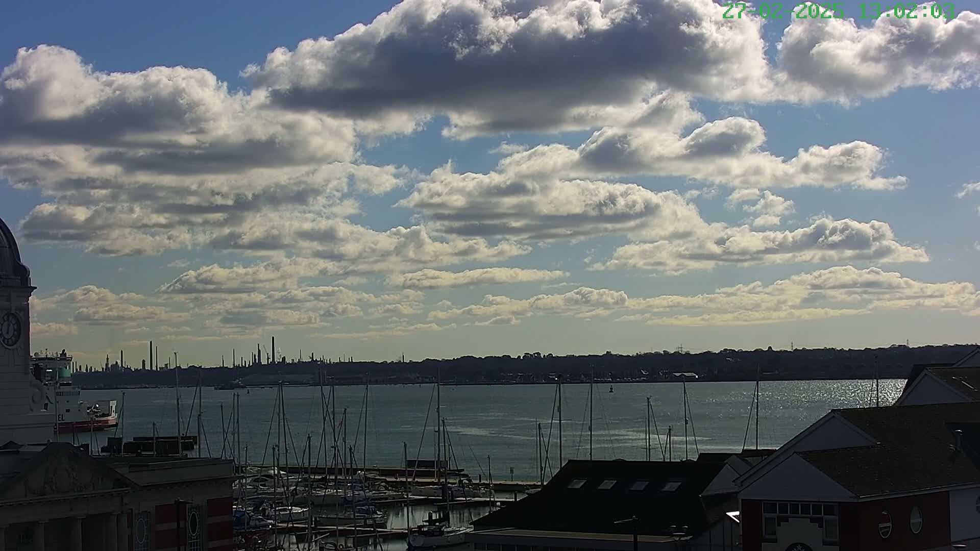 A partly cloudy day reveals a harbor filled with sailboats, a distant industrial skyline, and buildings along the waterfront.