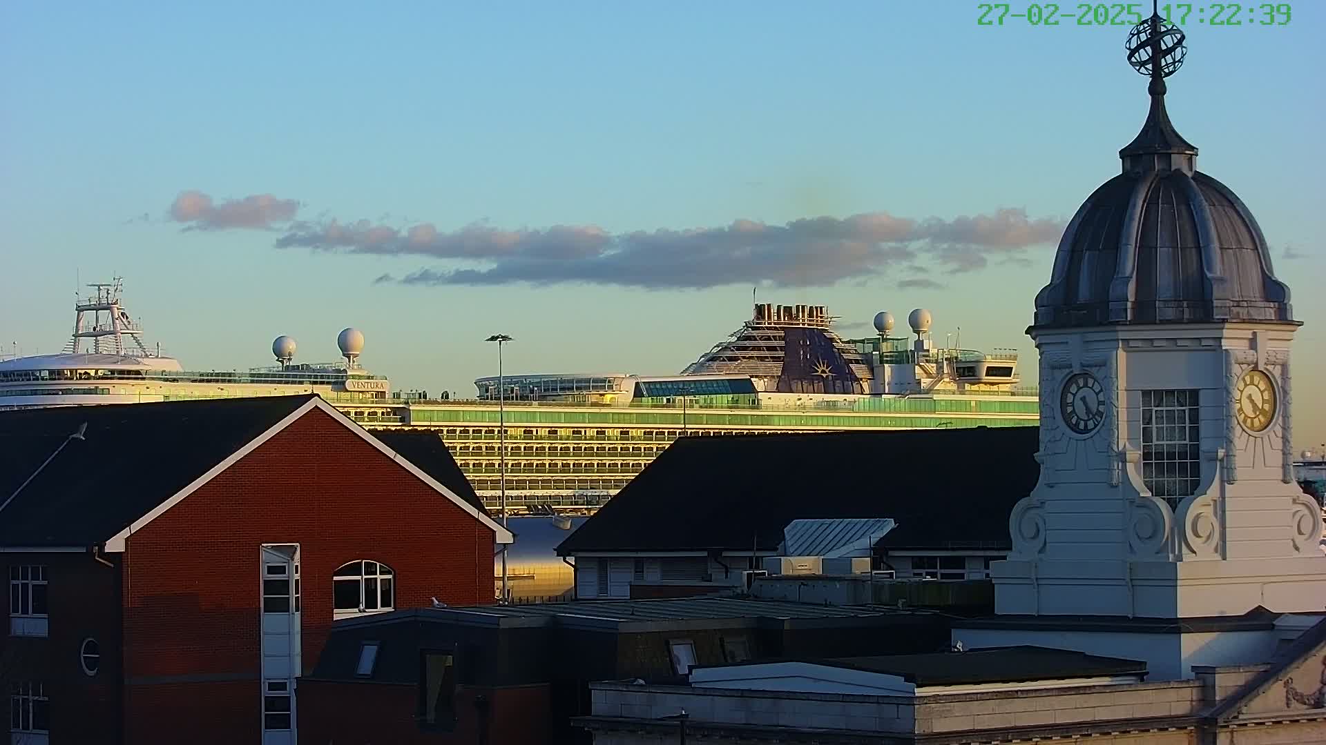 A large cruise ship is visible in the distance behind several brick buildings and a clock tower, under a mostly clear sky with a few clouds.