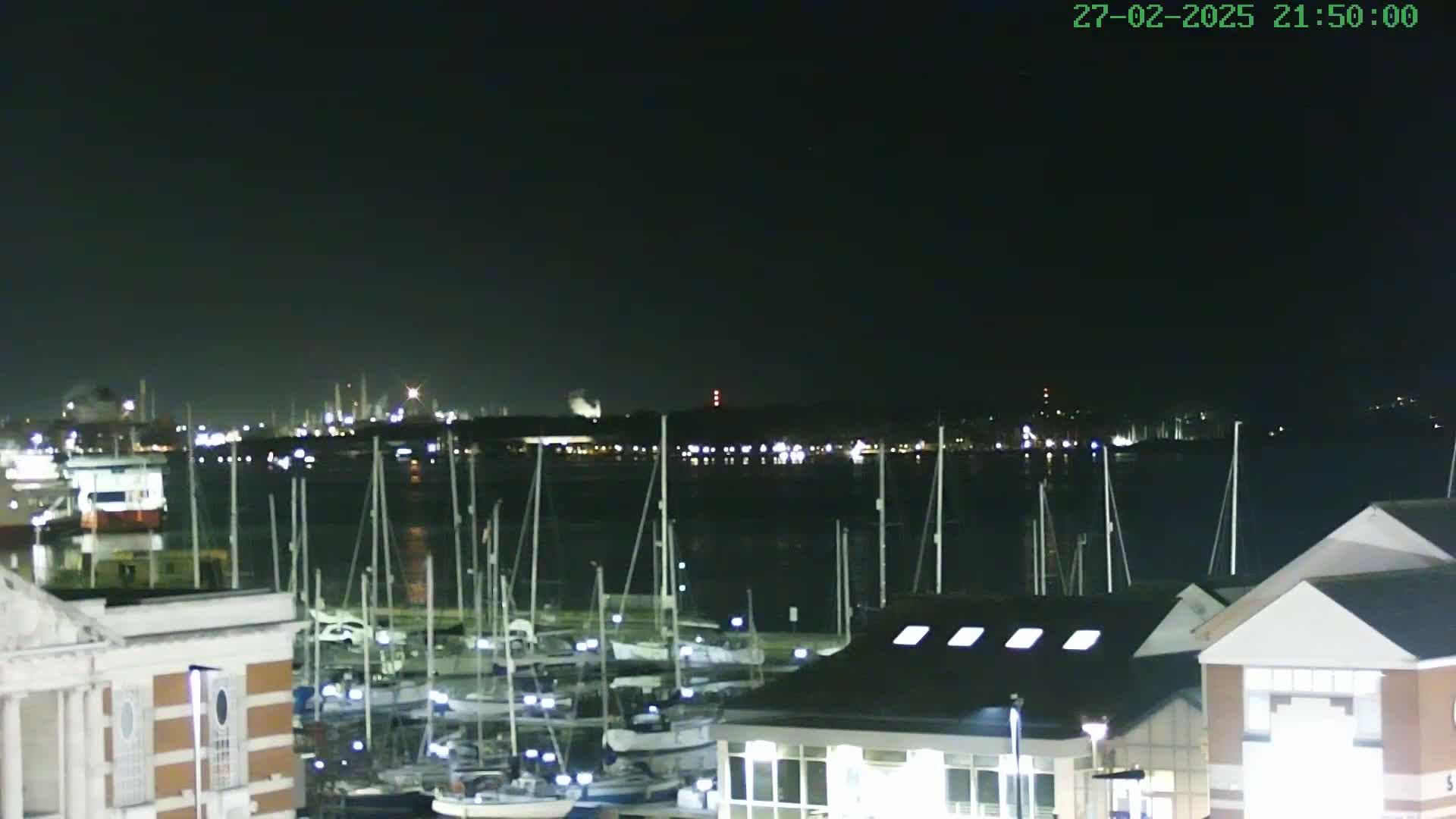 A nighttime view of a marina filled with sailboats, with a city skyline illuminated across the water under a clear, dark sky.