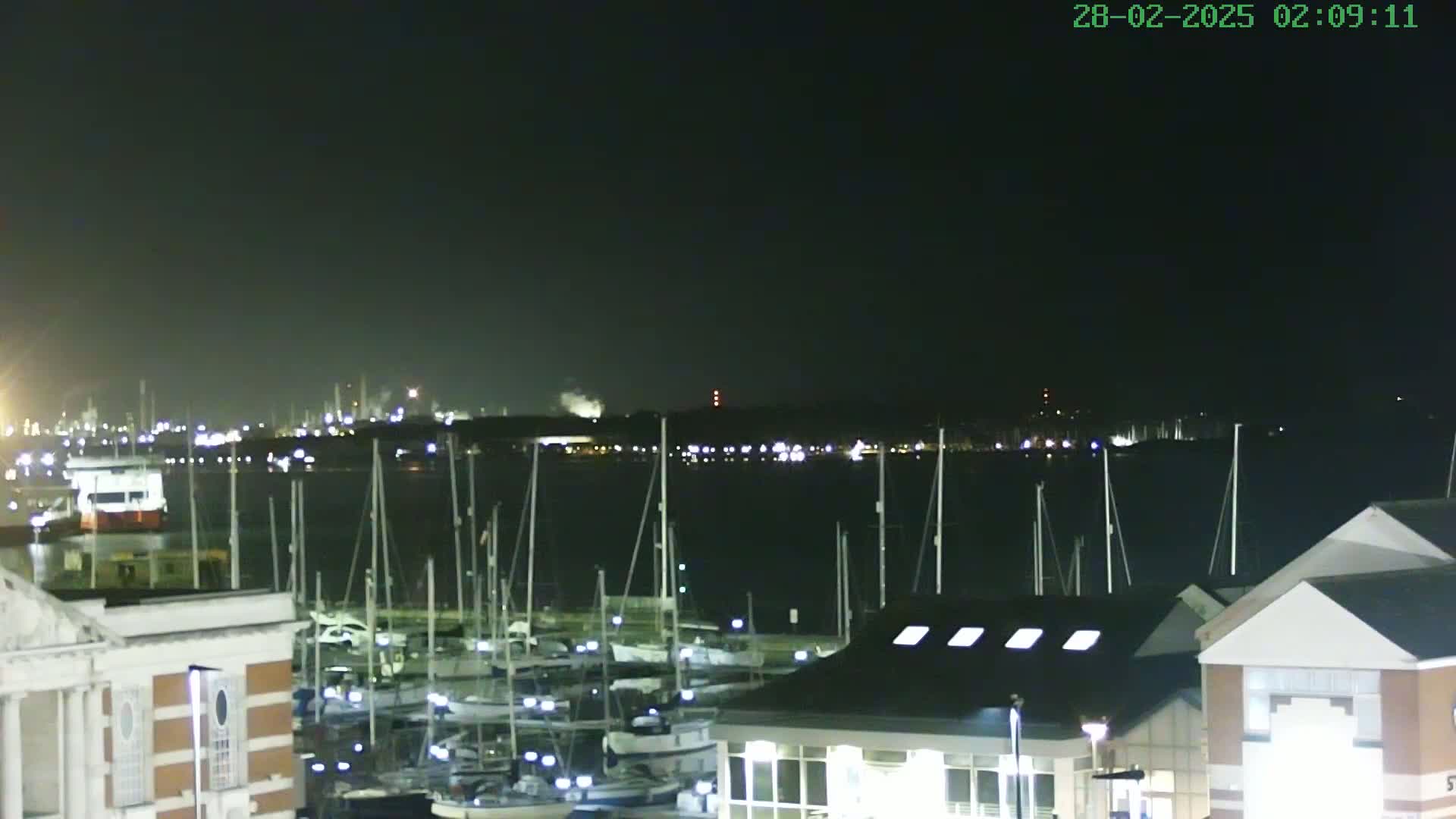 A nighttime view of a marina filled with sailboats, with a distant industrial shoreline faintly visible across the water under a dark, clear sky.