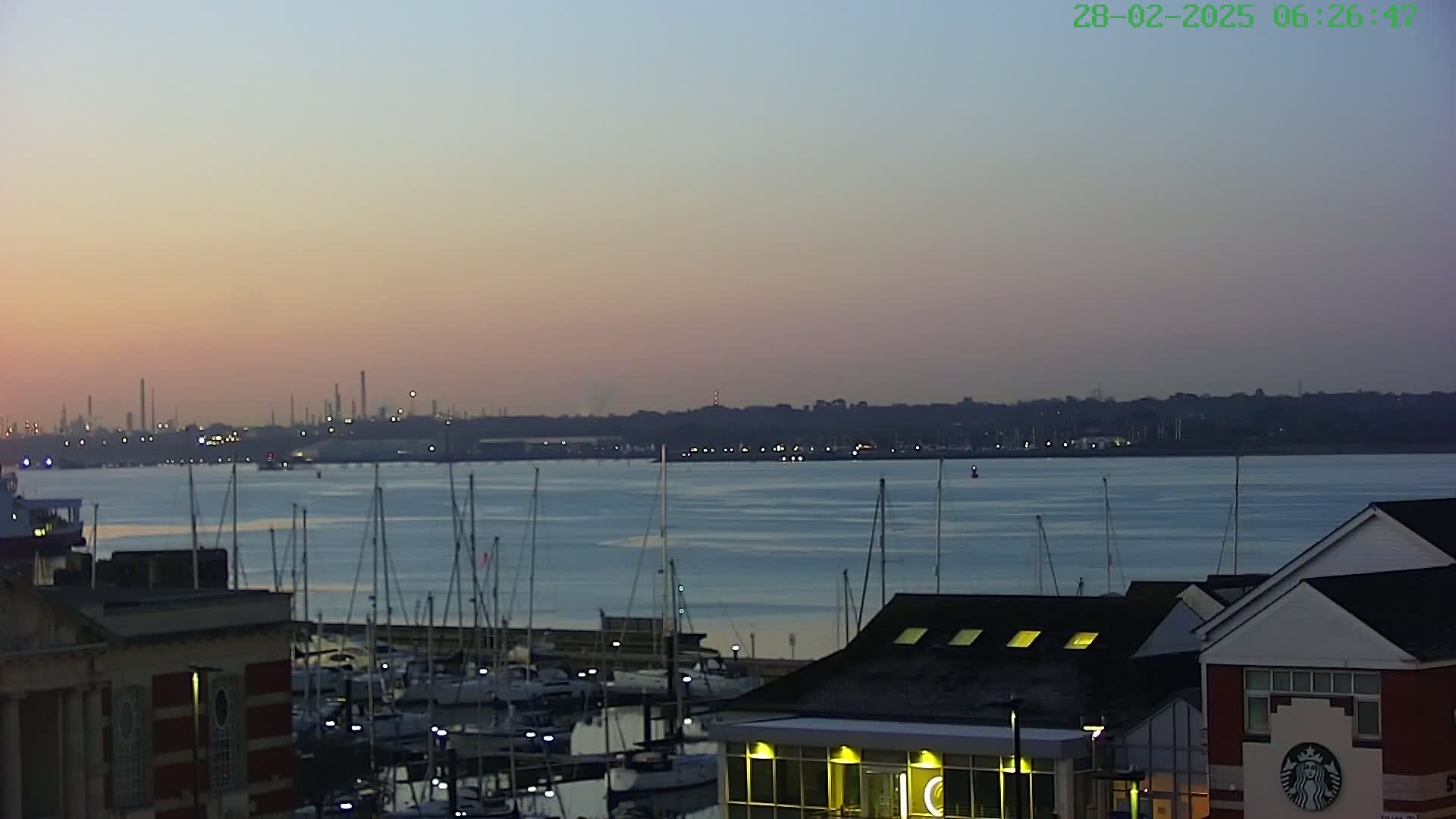 A calm body of water with many sailboats docked at a marina is seen at dawn, with an industrial area visible in the distance.