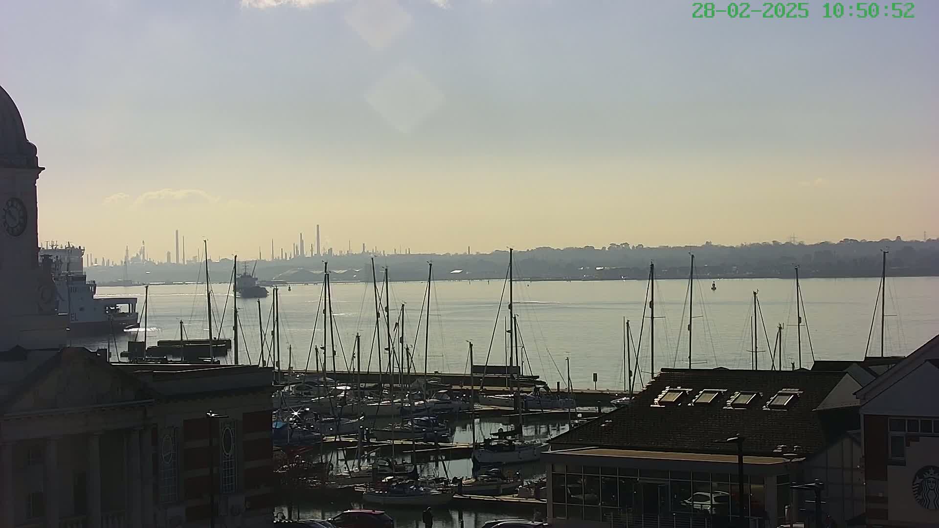 A harbor filled with sailboats is seen from an elevated vantage point, with a hazy industrial skyline visible across the calm water under a partly cloudy sky.