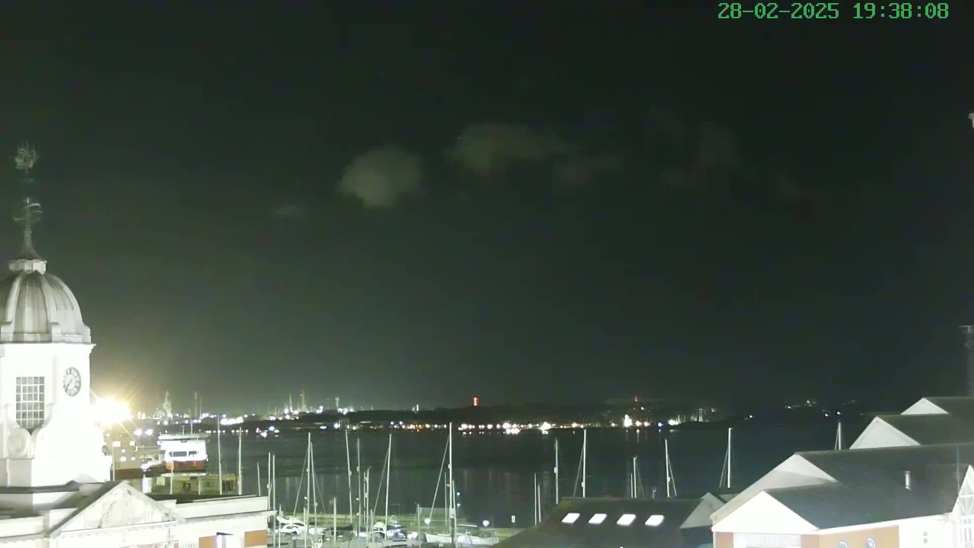 A nighttime view of a harbor with docked sailboats, a city skyline in the distance, and a clock tower in the foreground under a partly cloudy sky.