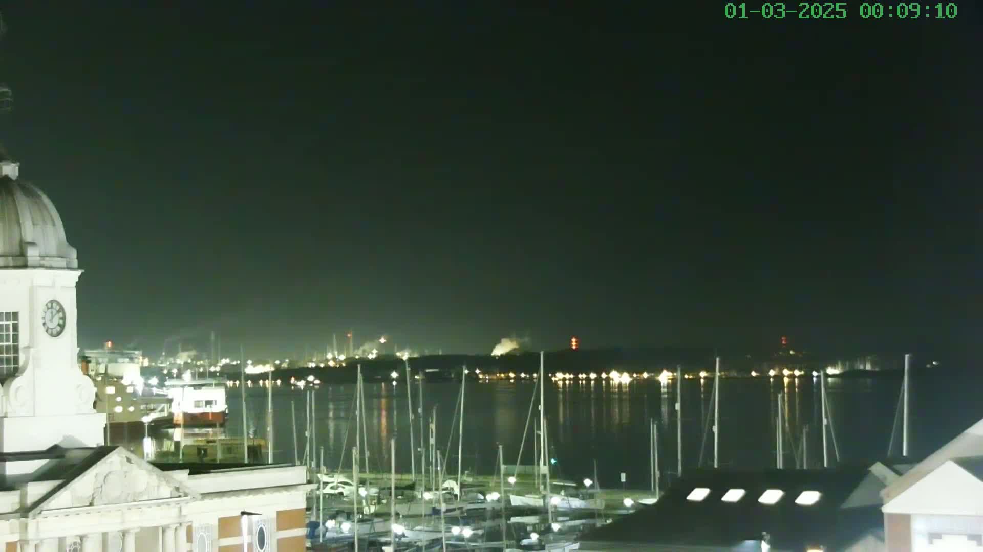 A nighttime view of a harbor filled with sailboats, with a clock tower and buildings in the foreground and industrial lights across the water.