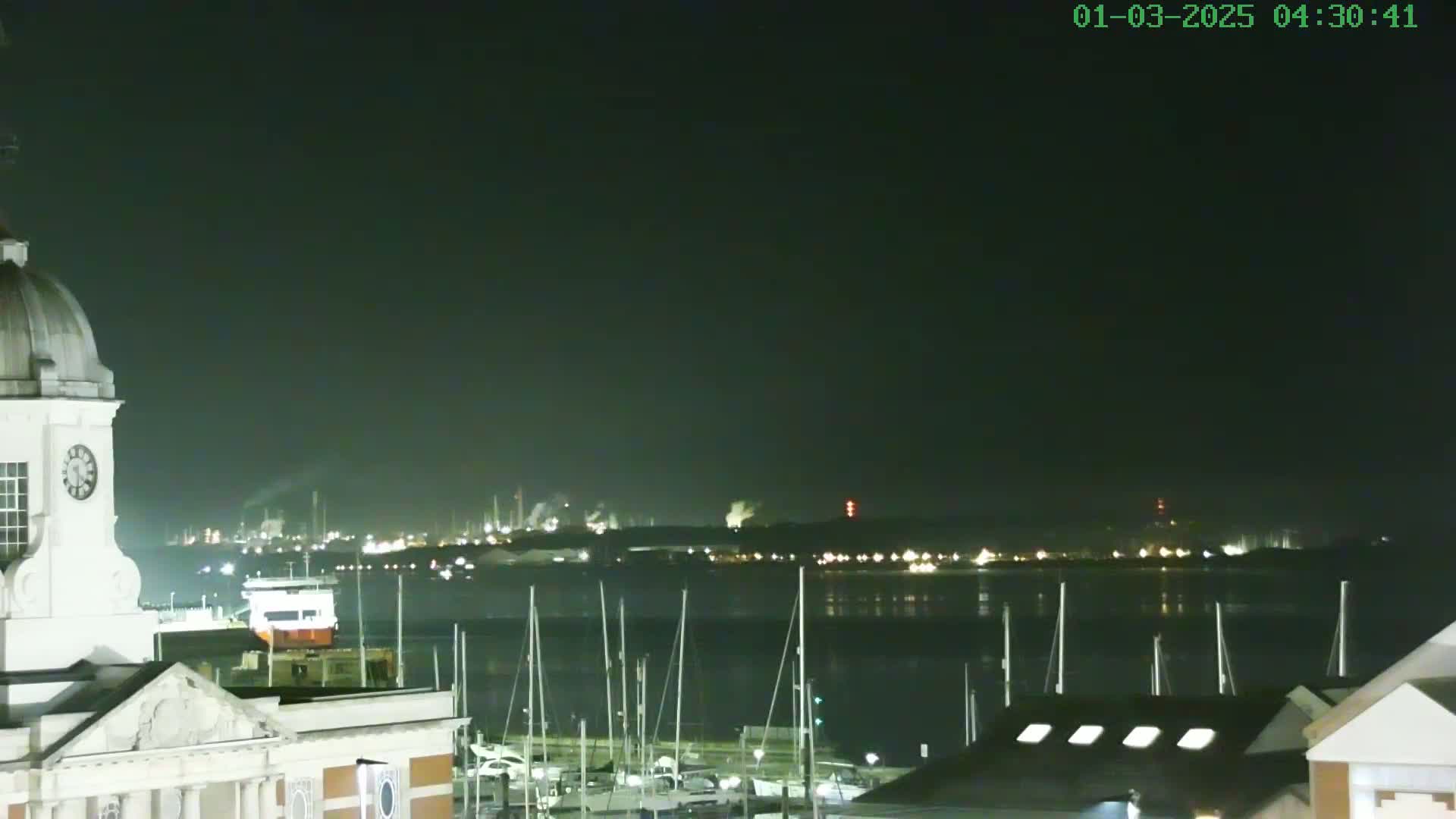 A nighttime view from atop a building shows a calm body of water with numerous masts of docked sailboats, an industrial area glowing in the distance, and a clock tower in the foreground.