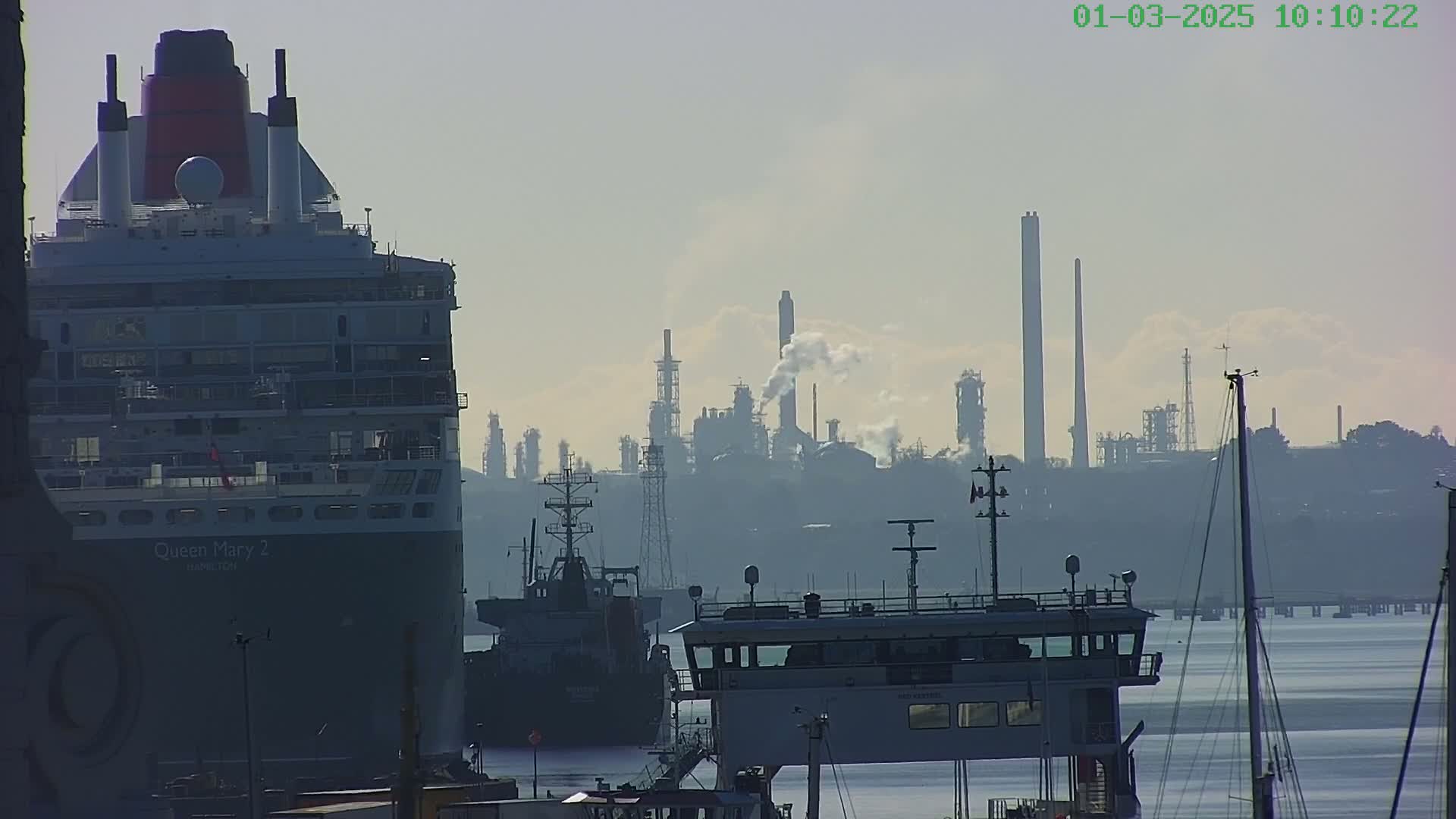 The stern of a large cruise ship is visible in the foreground, with an industrial refinery emitting smoke in the hazy background across a calm body of water.