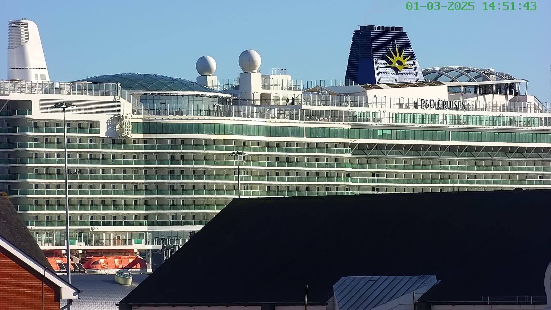 A large cruise ship with numerous decks and balconies is docked on a sunny day.
