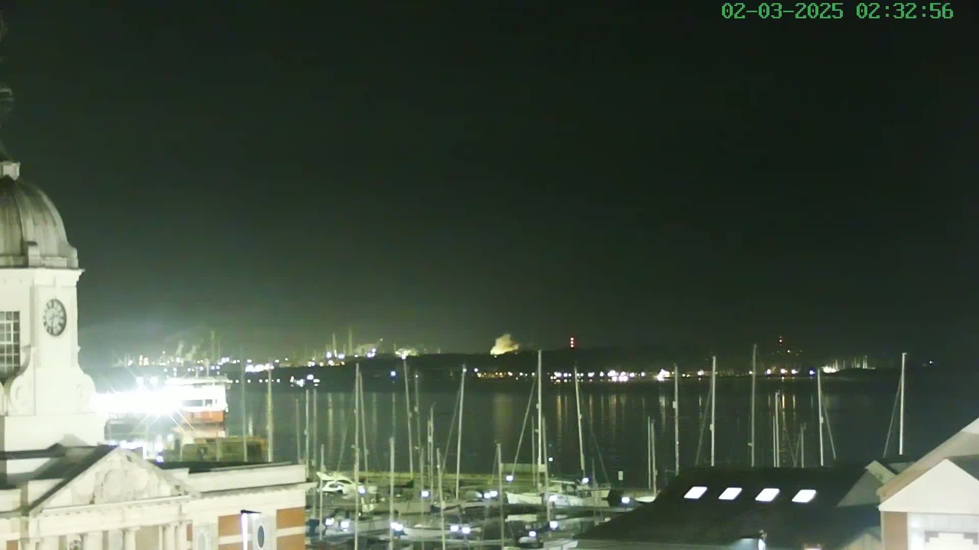 A nighttime view shows a harbor filled with docked sailboats, a distant illuminated industrial area, and a white building with a cupola in the foreground under a dark, clear sky.